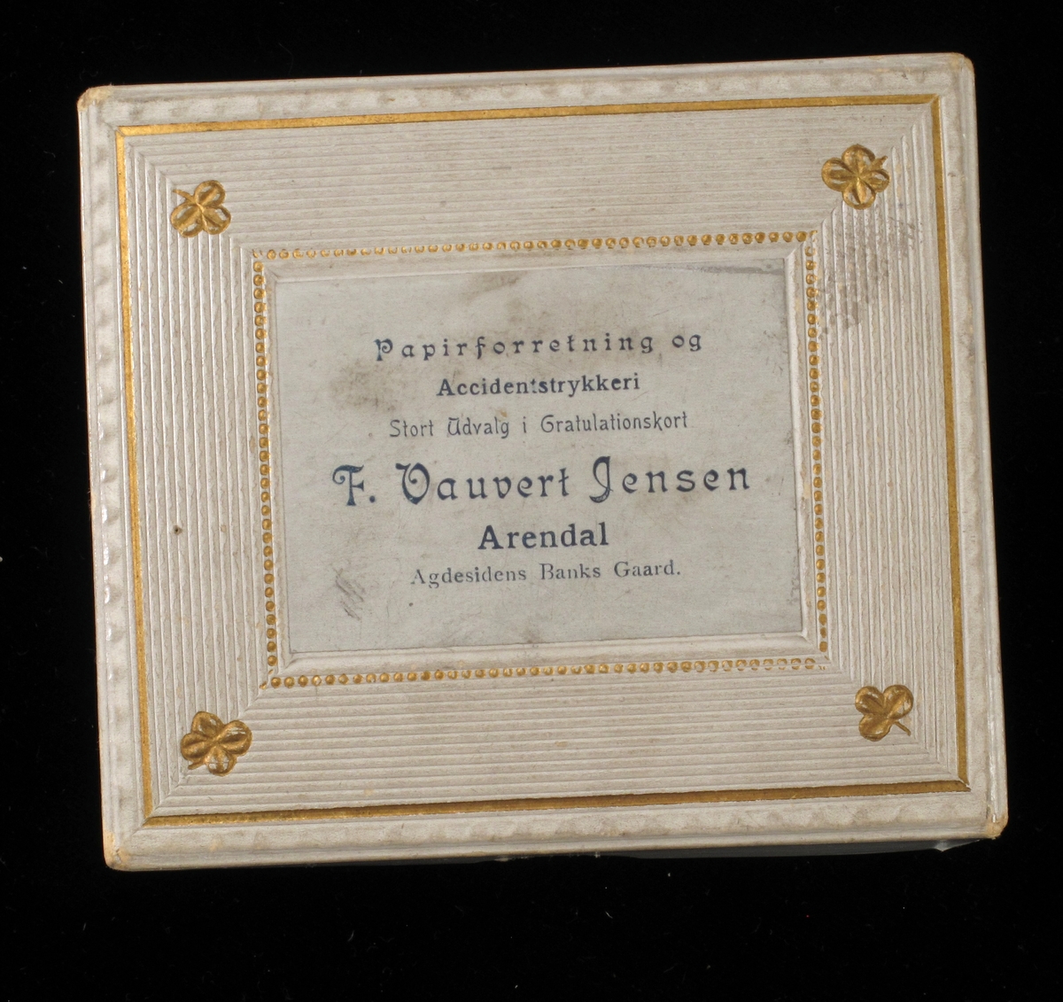 Vareeske av kartong, dekorert med påtrykt mønster og firmanavn. Eskelokket er dekorert med preget mønster, linjer, perler og firkløver.