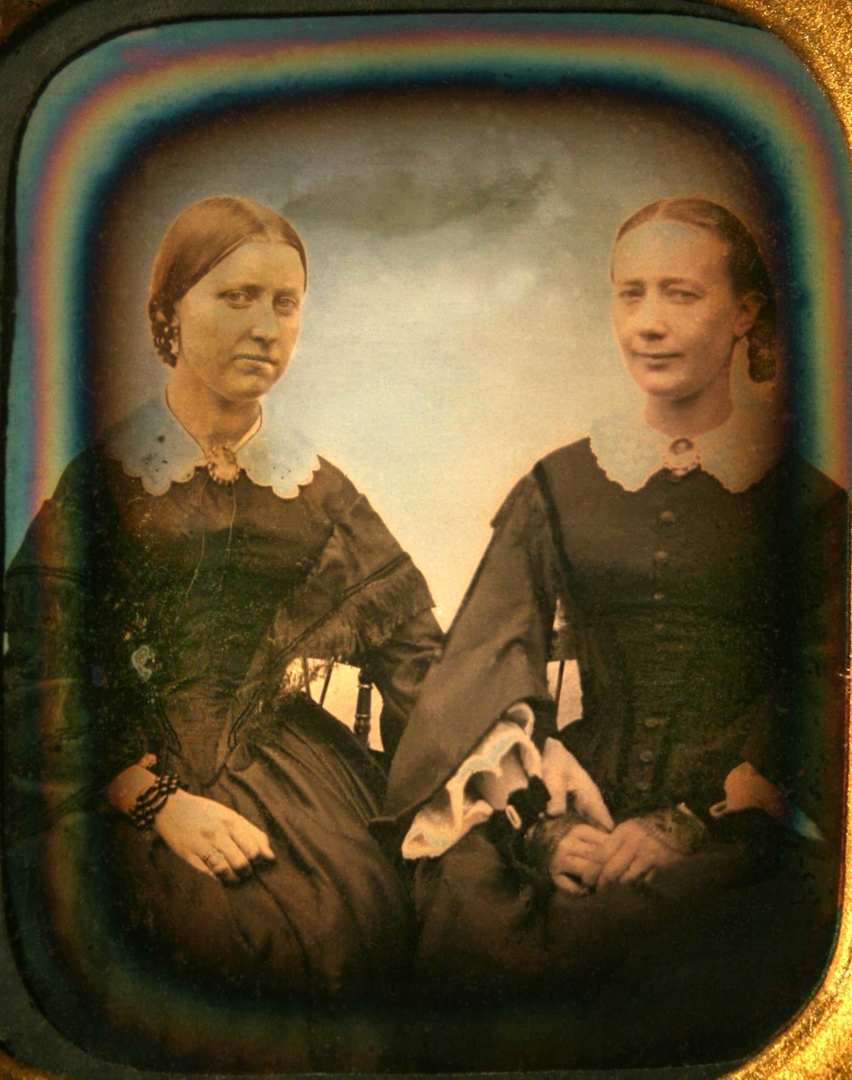 To sittende kvinner. Portrett av Laura Larssøn (f. 17/2 1830 i Bratteklev - d. 1908) og Edeline Anette Larssøn (f. på Torjusholmen 22/10 1834 - d.1915 på Merdø).   
Tv. sittende kvinne med glatt tilbakestrøket hår med midtskill, flette i nakken, mørk kjole som går ned i spiss midt foran, hvit krave, ermer med et frynset kast over. Hvit blondekrave med kamé.  
Th. sittende kvinne, glatt hår med midtskill, flette sees th., sort forknappet kjole med fløyelsbånd, hvit blondekrave med kamé, meget vide ermer med hvitt stoff i ermeåpningen. 
