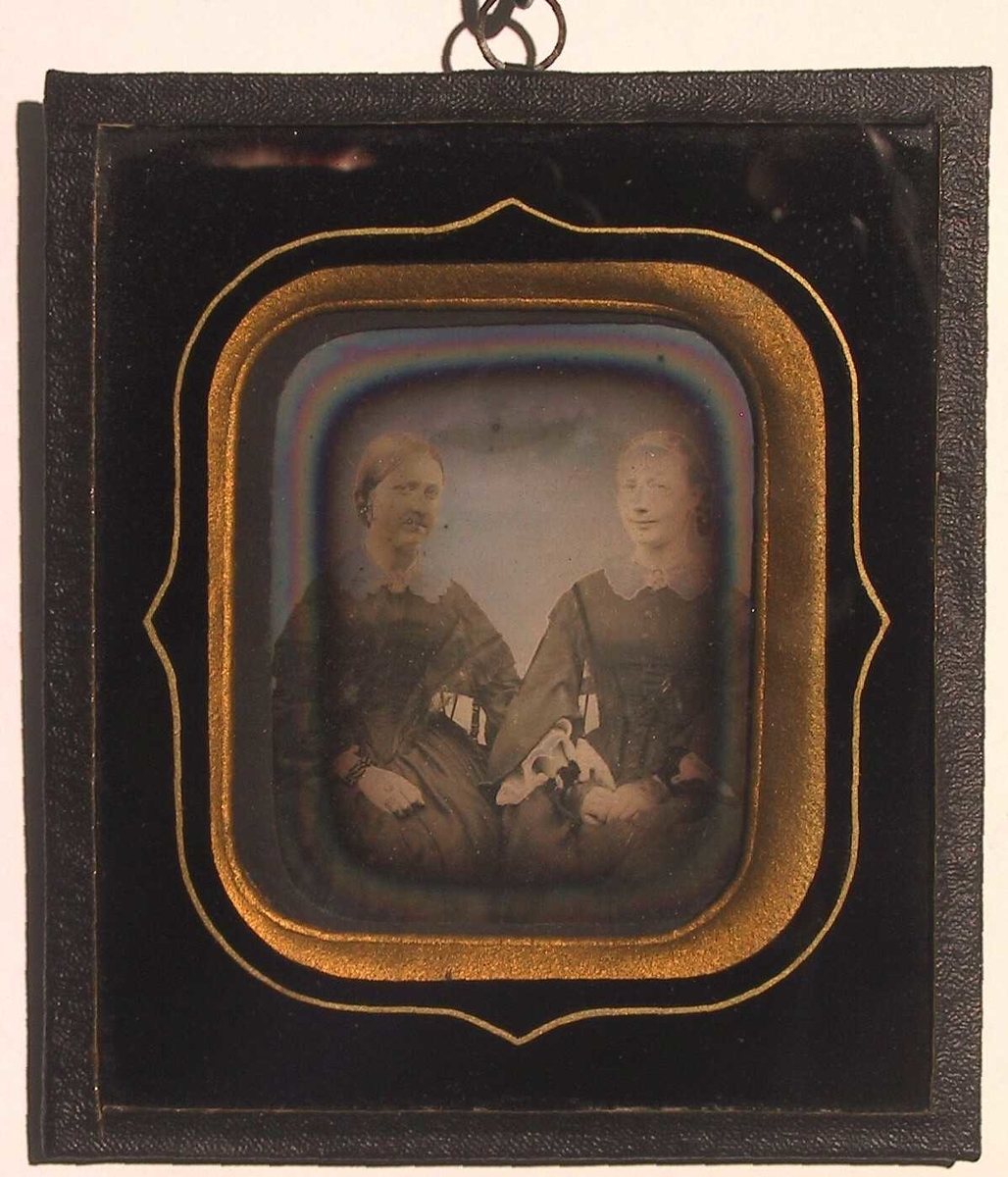 To sittende kvinner. Portrett av Laura Larssøn (f. 17/2 1830 i Bratteklev - d. 1908) og Edeline Anette Larssøn (f. på Torjusholmen 22/10 1834 - d.1915 på Merdø).   
Tv. sittende kvinne med glatt tilbakestrøket hår med midtskill, flette i nakken, mørk kjole som går ned i spiss midt foran, hvit krave, ermer med et frynset kast over. Hvit blondekrave med kamé.  
Th. sittende kvinne, glatt hår med midtskill, flette sees th., sort forknappet kjole med fløyelsbånd, hvit blondekrave med kamé, meget vide ermer med hvitt stoff i ermeåpningen. 