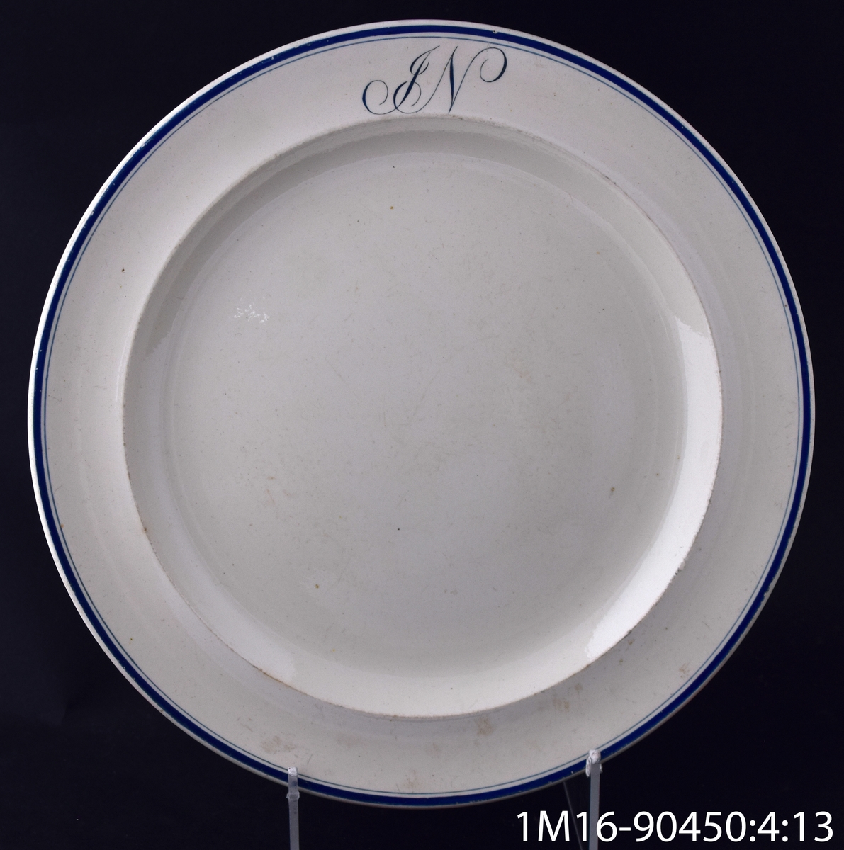 Tallrik, flat, av flintgods, med en bredare och en smalare blå rand längs kanten och initialerna JN, som står för John Nonnen. Tillverkare: Wedgewood.