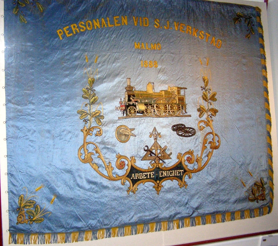 Rektangulär fackföreningsfana av blått broderat siden, för en järnvägsmannaförening i Malmö.
I mitten syns ångloket SJ Cb 339 Hervara, under detta ett bevingat hjul, en hjulaxel, samt ett arrangemang av verktyg och växtrankor.
Text i gult : "Personalen vid S.J. verkstad Malmö 1888 Arbete Enighet". Fanan är broderad i guld, svart och bruna nyanser med ett lok på mitten och eklöv i hörnen. Broderierna är utförda i stjälksöm och schattérsöm, vissa partier är även applicerade. Fanan har blågula fransar i kanterna.

Fanan är mycket blekt på den sidan som har legat uppåt i monter. Se sista bilden för jämförelse med undersidan.