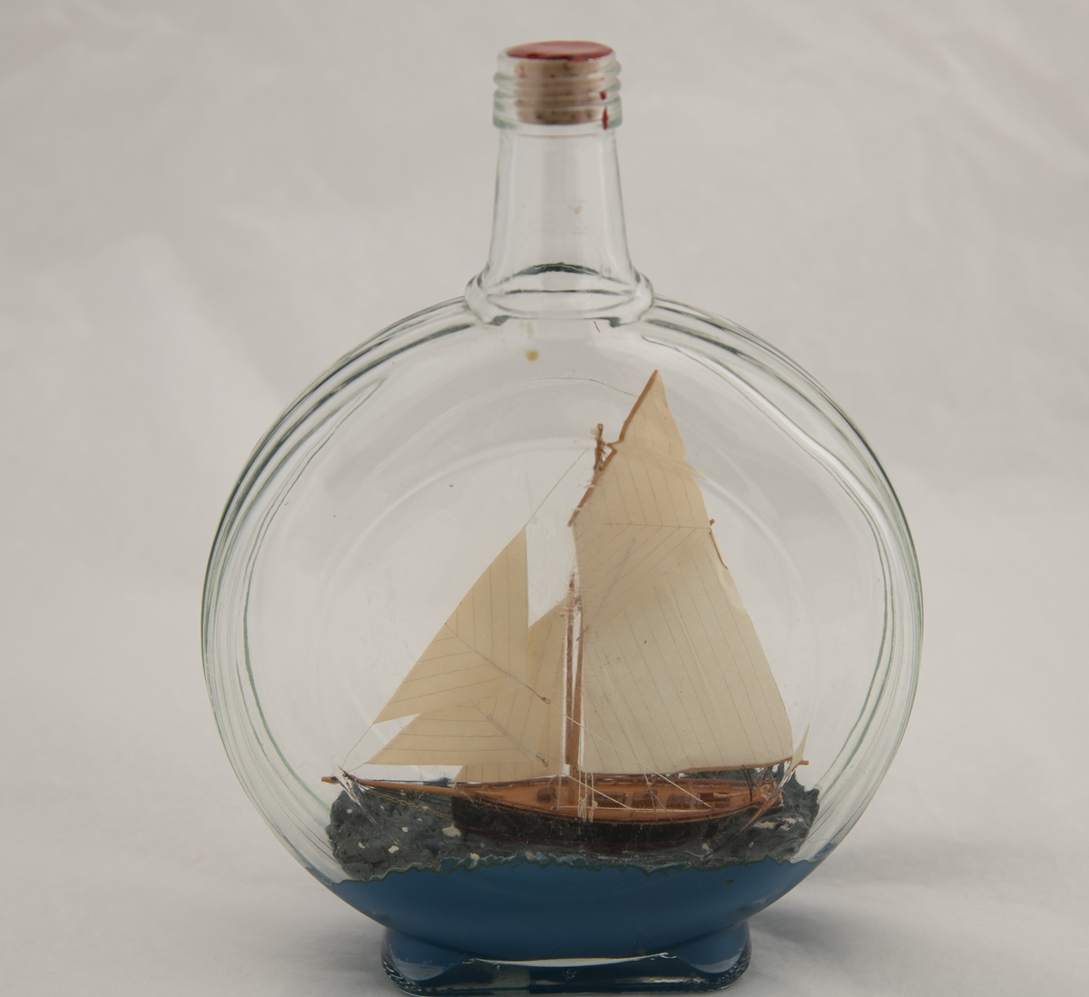 Flaske, klart glass. Lav kork med rød lakk. Smal rektangulær bunn, flatt skiveformet korpus, med riller langs den smale kanten og en ring på hver side (svakt forhøyd midtparti). Kort hals, skrugjenger. Ganske tykt lag med blå masse som bunn, sortmalt treskute med hvite seil, to nedganger, to skylights og åpent rom bak ved roret. Skuta må være satt sammen av enkelte deler inne i flasken.