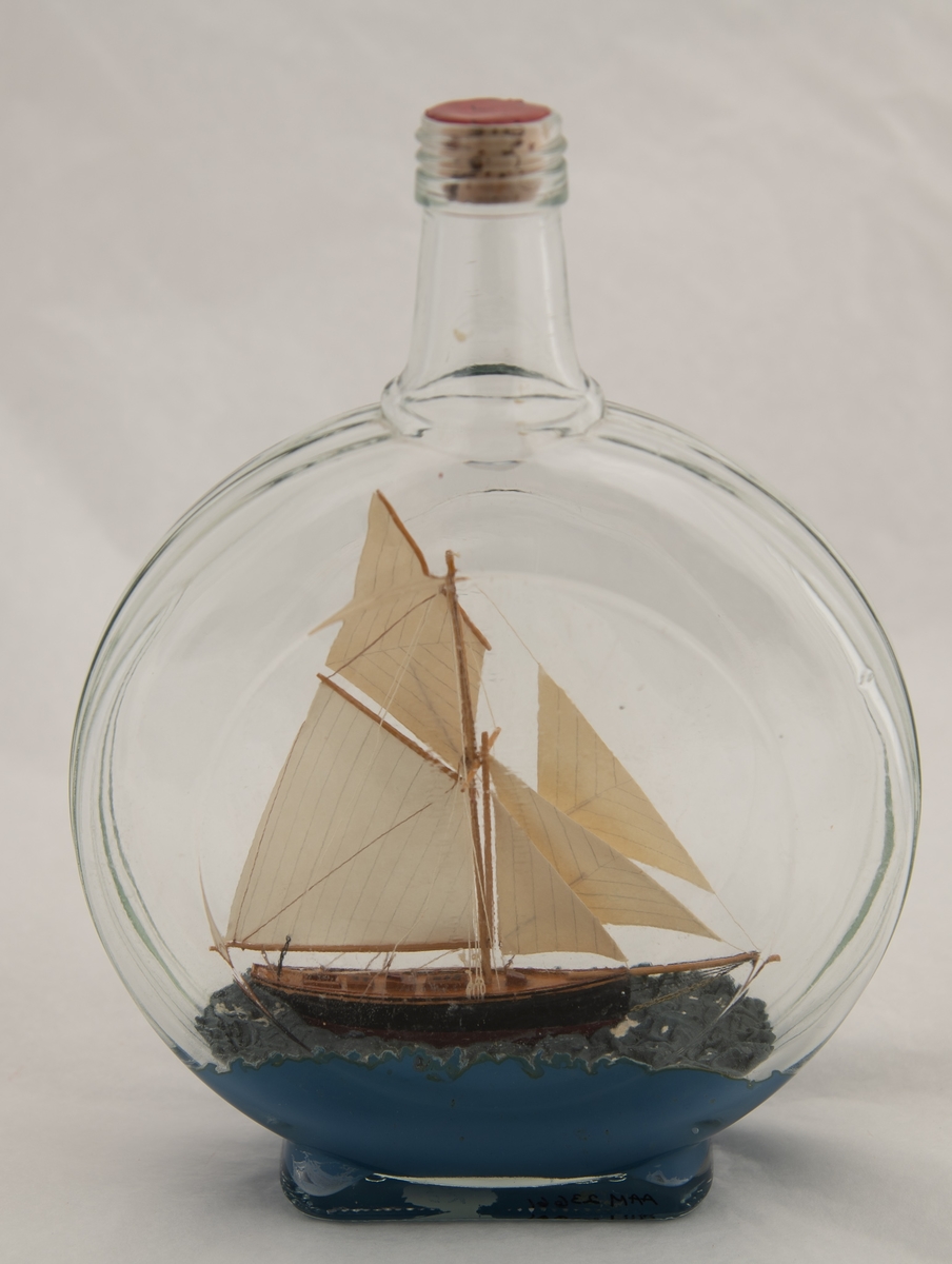 Flaske, klart glass. Lav kork med rød lakk. Smal rektangulær bunn, flatt skiveformet korpus, med riller langs den smale kanten og en ring på hver side (svakt forhøyd midtparti). Kort hals, skrugjenger. Ganske tykt lag med blå masse som bunn, sortmalt treskute med hvite seil, to nedganger, to skylights og åpent rom bak ved roret. Skuta må være satt sammen av enkelte deler inne i flasken.