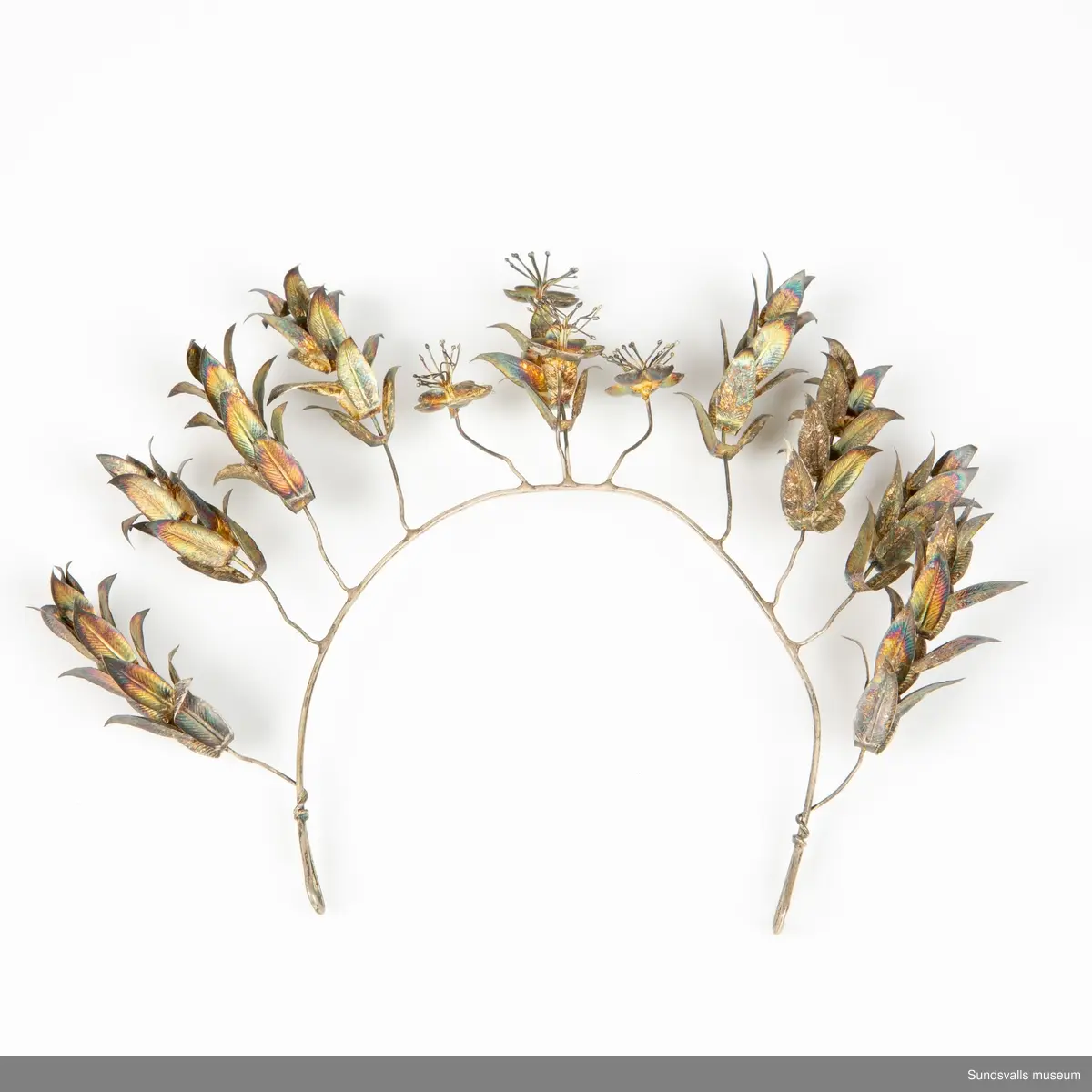 Diadem i böjd metalltråd med en ögla i var ände. Dekoration i form av metalltrådar med blommor och blad är fästade runt den böjda tråden.