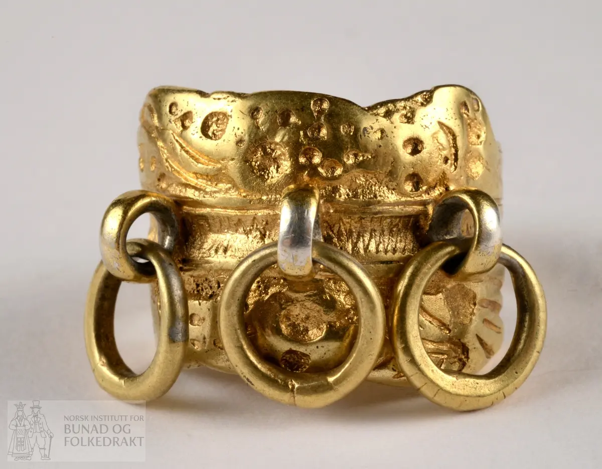Fingerring av sylv. Forgylt. Støypt med mønster i slangeform som går 2,5 ganger rundt ringen. Trambulert dekor midt på bandet/ slangen. 3 kraftige ringar påsett sringar av tråd midt framme. Ringen er gjort mindre ved å sette på ein nær flatvalsa tråd inne i ringen framme. 4 stempel, vanskelege å lese.