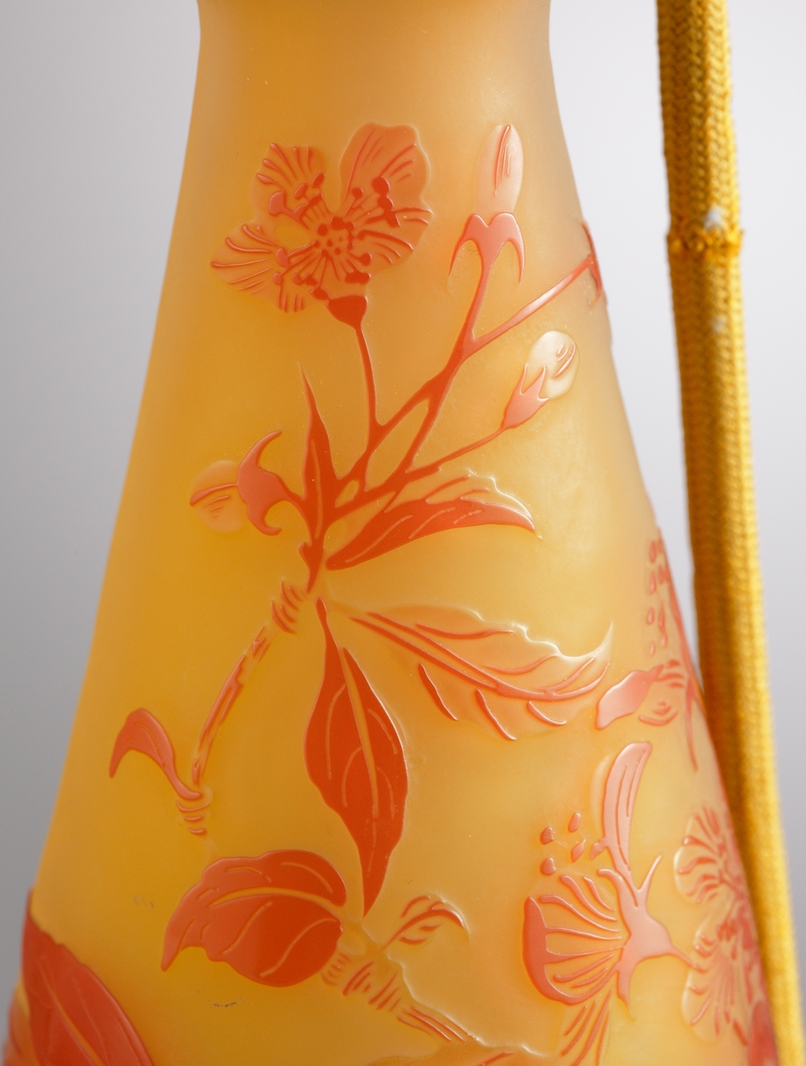 Luftfräschare i opakt gult glas med rött etsat överfång. Dekorens motiv är kvistar med blad och blommor. Överst på flaskan sitter en metallfattning med en textilslang försedd med en gummipuff.