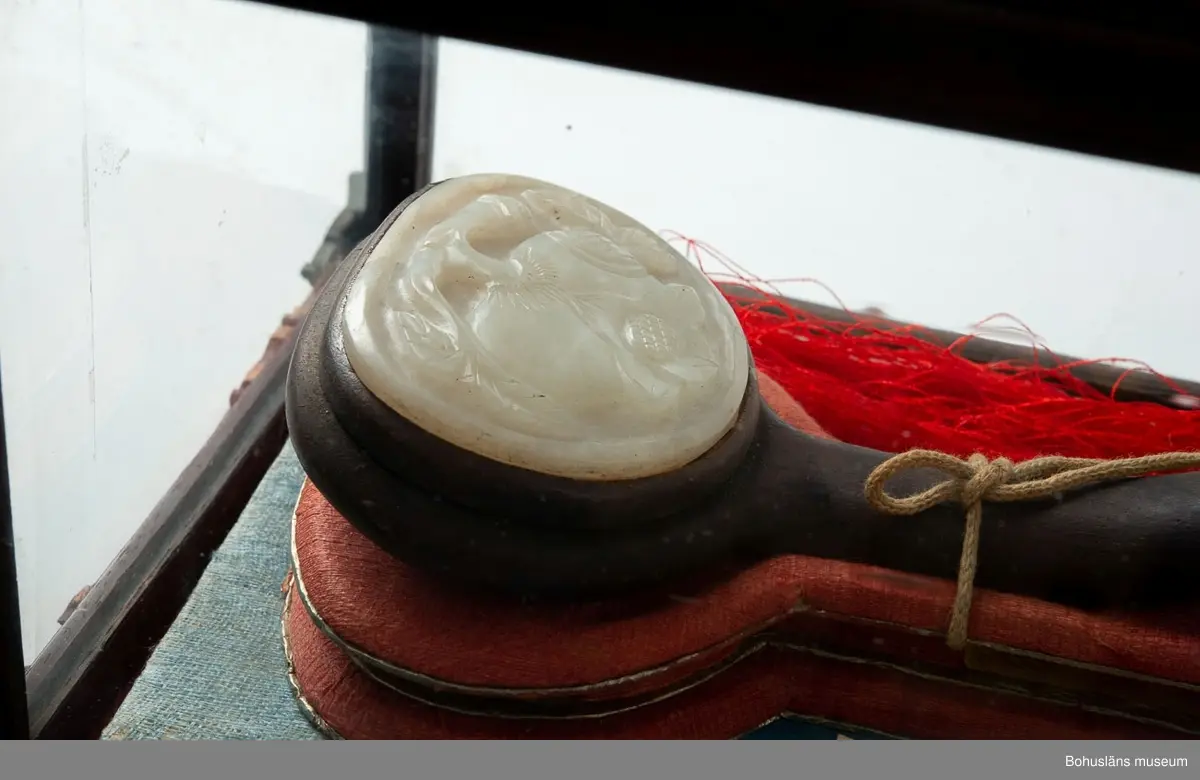 Tre stenar med ristade motiv i låda med glas.
Stenarna är troligtvis av mineralet nefrit som är besläktat med jade. Stenarna är infattade i ett träskaft och vilar på en textil.

Ur handskrivna katalogen 1957-1958:
"sarij", agatlikn. stenar o låda m. glas.
Lådans. L: 49,5 Br. 18 H: 26,5. Innehåll 3 agatliknande stenar  med djurornament, en tofs. Relativt välbevarat 18

Anteckning i 1872 – 1932 Gåfvor till Uddevalla museum:
År 1910
Mars
Ingenjör Johannes Johansson från Garn i Forshälla socken Bohusl. som vistats 23 år i Australien o på sistone i China, har skänkt Uddevalla museum 1) Ett riktigt jättestort siden-parasoll, siradt med blommor och figurer. [UM000683].
2) Ett par kinesiska flaggor i vimpelform av siden o med drakemblemet insyddt. [UM000684, UM000685].
3) trenne i en slags mahogny-glaslåda förvarade agatliknande stenar med konstnärligt ristade gravyrer. [UM000647].
Parasollen kallas på kinesiska ”Sutja” och Agatlådan ”Sarij”. Båda äro avsedda att vi fästliga tillfällen bäras framför ägaren såsom tecken till, att dessa äro av hög samhällsställning. De tilldelas i regel endast mandariner eller i övrigt särskildt betrodda. Vid avresan från Kina, där Johannes Johansson varit arbetsledare fick han av sina järnvägsarbetare i befolkningen i det distrikt, där järnvägen byggdes under honom såsom chef mottaga de nämnda dyrbara gåvorna såsom ett vittnesbörd om, huru uppburen han i egenskap av byggnadschef varit.

Ur handskriven katalog över Uddevalla - Musei Historiska - samlingar: Utländska föremål = De etnografiska föremålen upprättad  år 1916 av Knut Adrian Andersson, Intendent från 1 Oktober 1915:
No 24. "Sarij" kinesiskt namn på graverade (3 stycken) agat-liknande stenar med konstnärliga gravyrer; förvarade i tillhörande mahogni?-låda med glas.
Hedersgåva från Kineser till Herr Johannes Johansson i Garn Forshälla socken för hans arbete i Kinas tjänst. Skänkt av honom 1910 till Uddevalla museum.

Enligt uppgift i nedanstående artikel hade Johannes Johansson vistats 23 år i främmande land. Han hade skött järnvägsbyggen i Australien, och därefter lett byggandet av järnvägen mellan Nanking och Shanghai i Kina.
Uppgift i "Sveriges befolkning 1890" tyder på att Johannes Johansson var född 1865 och hade titeln sjöman.

Lappkatalog: 10

Litteratur: 
Artikel "Exotica" i Bohusläningen den 14 augusti 1941.