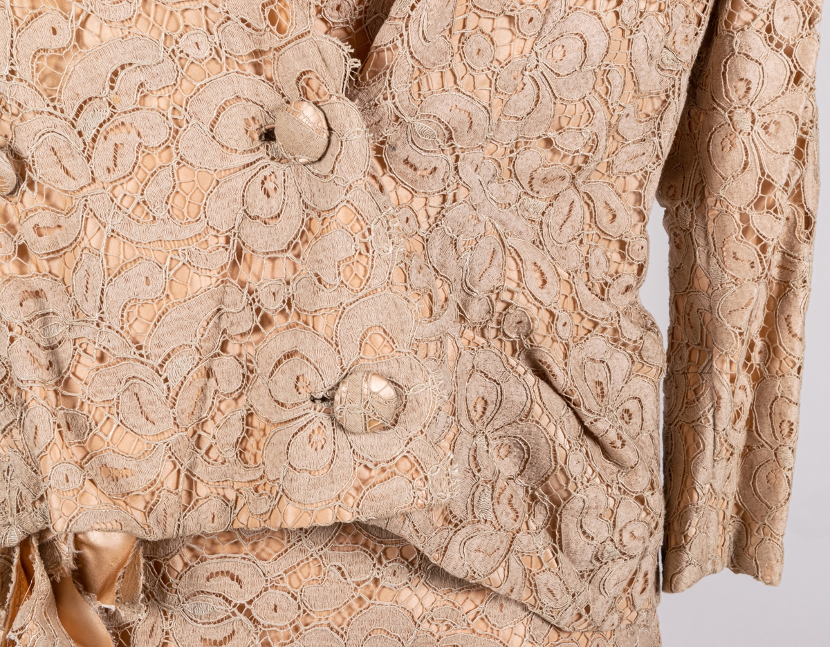 Tvådelad klänning (jacka och kjol). Beige, bomullsspets på gulrosa foder i duschess. Kort jacka med 3/4 lång kimonoskuren ärm. 2 stolpfickor fram. Kraglös med spestkanten som kantavslut. Omlottknäppning med 3 tygklädda knappar. Rak kjol med omlott fram. Dragkedja bak, knäppt med hyska och hake. Ewe-modell. Konfektion.
