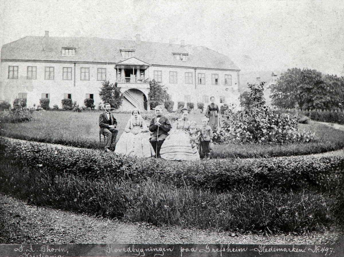 Ringsaker, Nes, Grefsheim gård, hovedbygningen, gruppe familien Egeberg. Foto av sorenskriver Johan Egeberg Mellbye (1804-1894) i midten med hustru Johanne (Hanna) Jacobsdatter Meyer. Ove Mellbye med hustru Sophie Munch, barn Johan Egeberg Mellbye (1866-) og Sofie Christine Mellbye (1868-). Egeberg overtok gården i 1867.  
Tittel: "Hovedbygningen paa Grefsheim Hedemarken" 
Fotograf P. A. Thorén nr 497