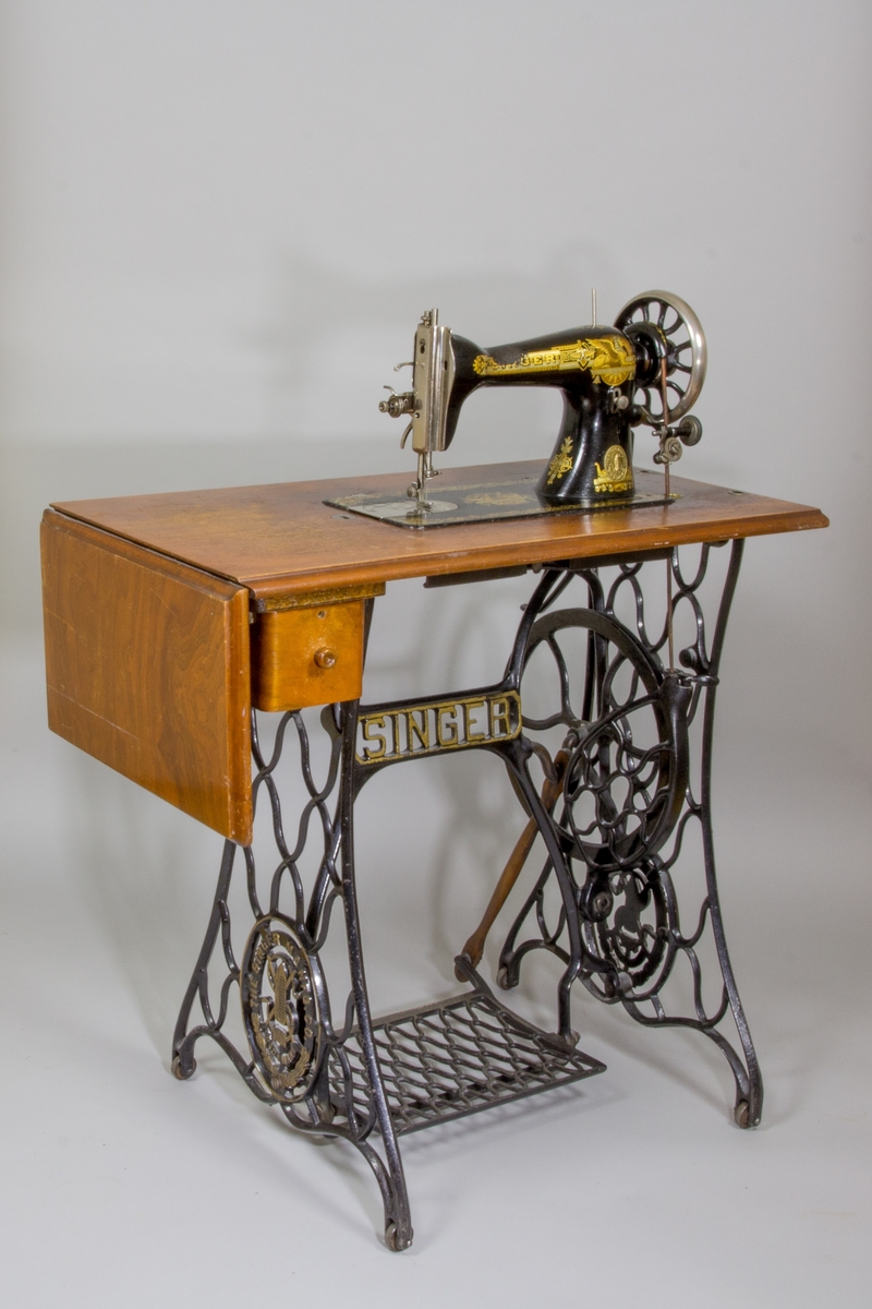 Symaskin, trampsymaskin,  på bord av märket Singer. Maskinen svartlackerad med dekorativt guldtryck i antikiserande stil samt text ”Singer”. Förnicklade detaljer. Skivan av trä med en fällbar del och en låda. Benställning och fottramp av svartlackerat gjutjärn. Trähuv med guldornament och texten ”Singer Manufg. c.o. Trade mark ”.