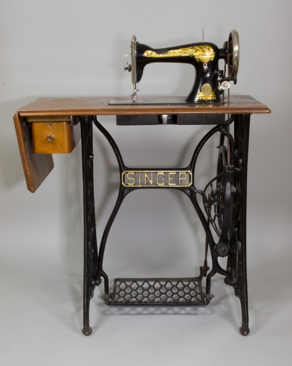 Symaskin, trampsymaskin,  på bord av märket Singer. Maskinen svartlackerad med dekorativt guldtryck i antikiserande stil samt text ”Singer”. Förnicklade detaljer. Skivan av trä med en fällbar del och en låda. Benställning och fottramp av svartlackerat gjutjärn. Trähuv med guldornament och texten ”Singer Manufg. c.o. Trade mark ”.