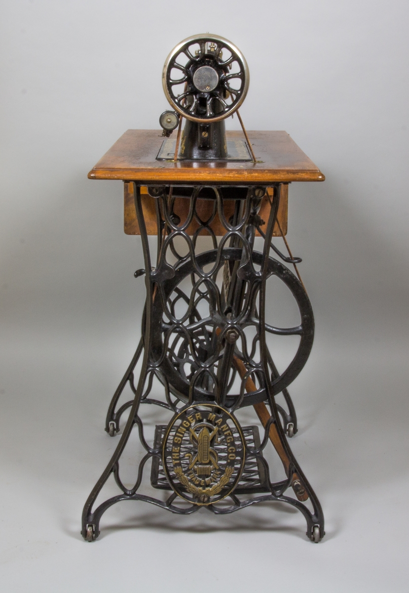 Symaskin, trampsymaskin,  på bord av märket Singer. Maskinen svartlackerad med dekorativt guldtryck i antikiserande stil samt text ”Singer”. Förnicklade detaljer. Skivan av trä med en fällbar del och en låda. Benställning och fottramp av svartlackerat gjutjärn. Trähuv med guldornament och texten ”Singer Manufg. c.o. Trade mark ”.