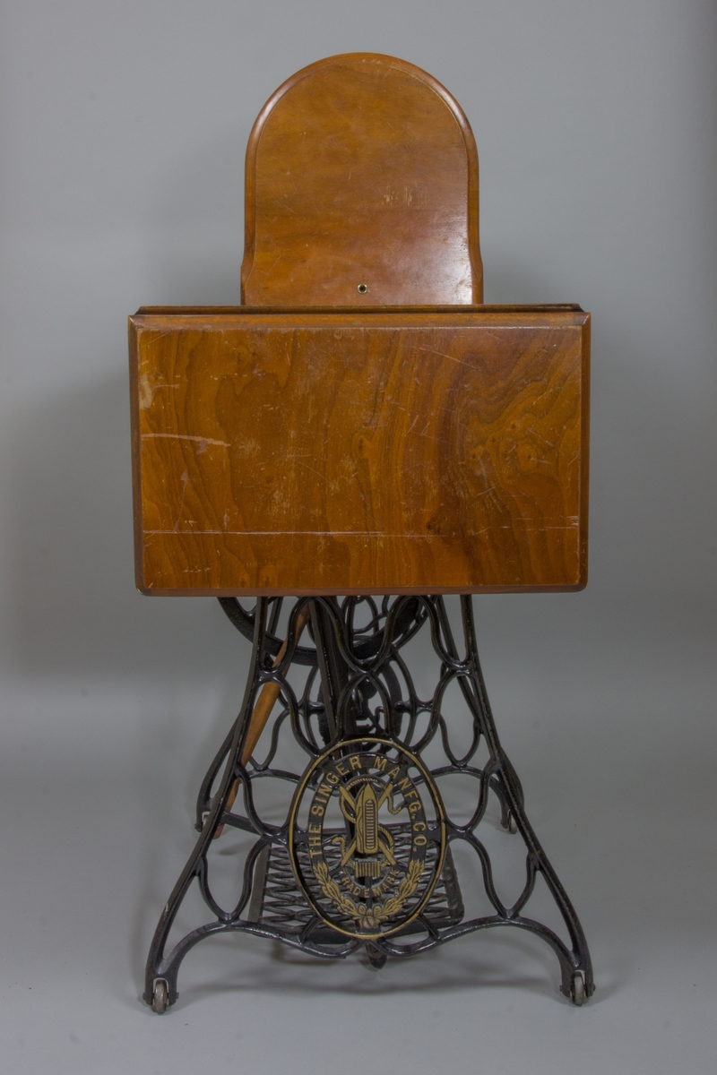 Symaskin, trampsymaskin,  på bord av märket Singer. Maskinen svartlackerad med dekorativt guldtryck i antikiserande stil samt text ”Singer”. Förnicklade detaljer. Skivan av trä med en fällbar del och en låda. Benställning och fottramp av svartlackerat gjutjärn. Trähuv med guldornament och texten ”Singer Manufg. c.o. Trade mark ”.