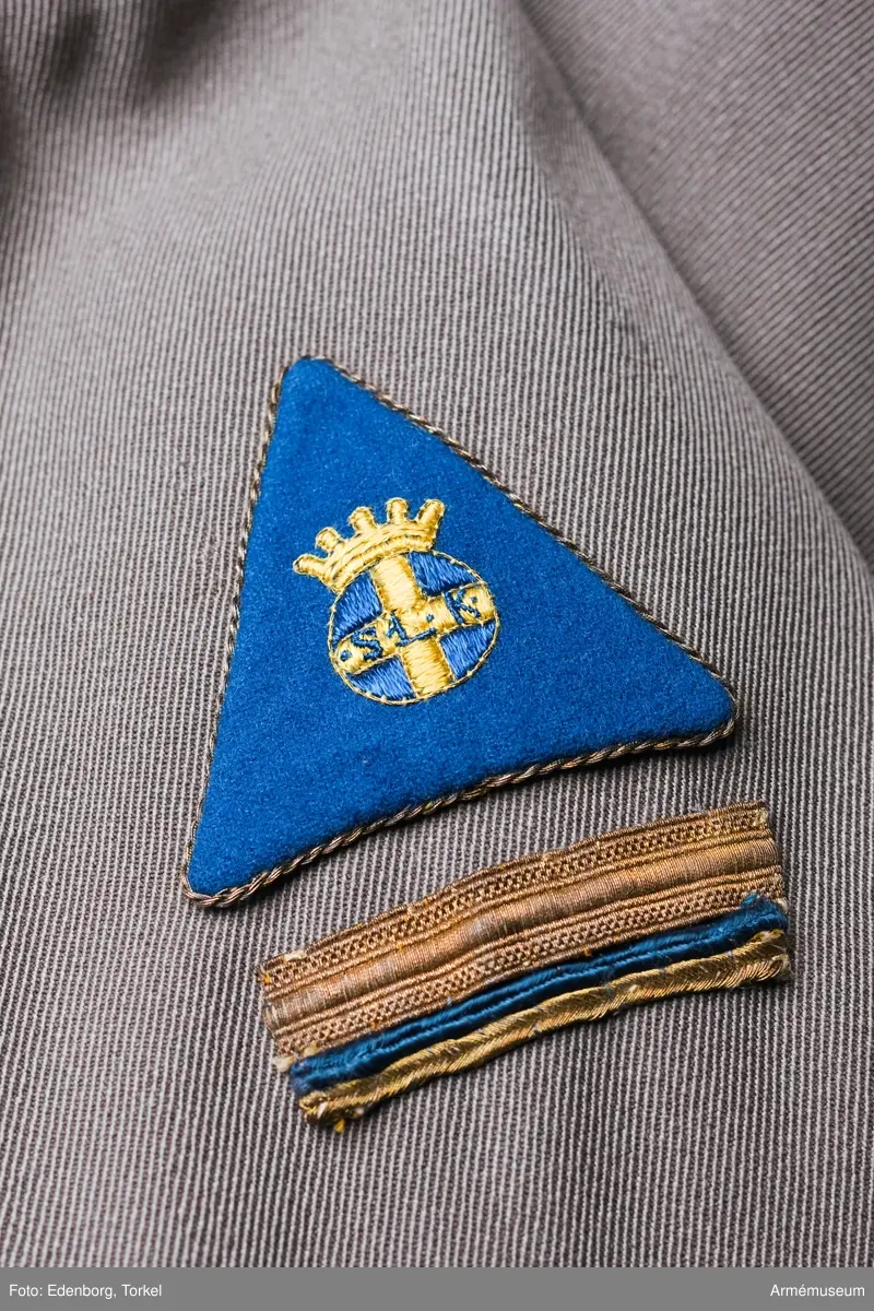 Stl 40. Ylleklänning knäppt till midjan med elva knappar (bakelit?). Ärmen blusande mot manschetten som knäpps med två knappar och  är 70 mm bred. Avskuren i midjan och sydd med nypveck mot midjesömmen. På livet ett smalt motveck, nedstickat från axeln till midjan. Söm mitt bak och blixtlås i vänster sida.
Skärp fäst i hällor och knäppt med två knappar. Två fickor, som  knäppes med knapp och är påstickade. Motveck mitt under var ficka och slutande strax nedanför dessa. Mitt bak ett efterveck.
På vänster ärm kårchefstecken: en bred guldgalon, en blå smal silkegalon och en smal guldgalon. Över detta tecken är fäst en rektangel av blått ylle, kantad med guldsnodd. Mitt på märket lottaemblemet broderat i gult/blått silke. Triangeln  betecknar flyglottor vars förening A Rudebeck var med och startade. Vit påknäppbar pikékrage.