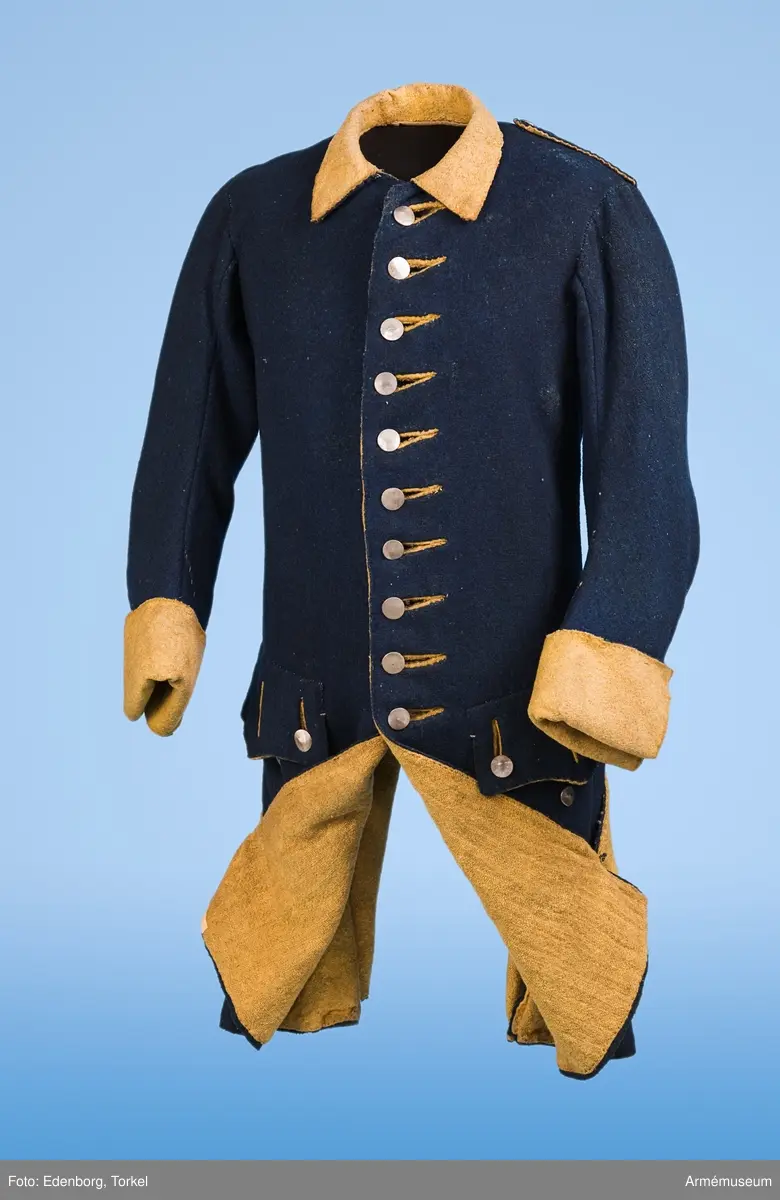 Rock av blått kläde med gul krage och ärmuppslag. Fodrad med gul boj i livet och med linne i ärmarna. Försedd med tennknappar.