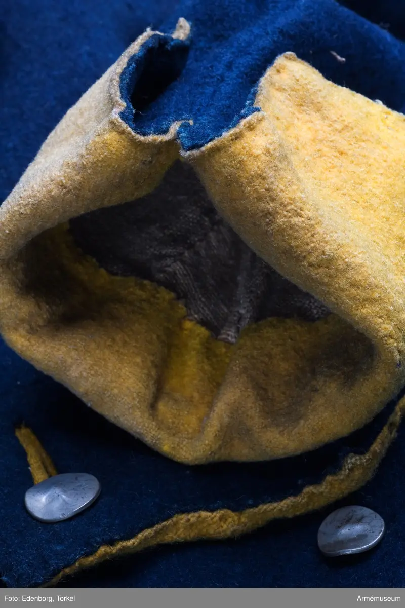 Rock av blått kläde med gul krage och ärmuppslag. Fodrad med gul boj i livet och med linne i ärmarna. Försedd med tennknappar.