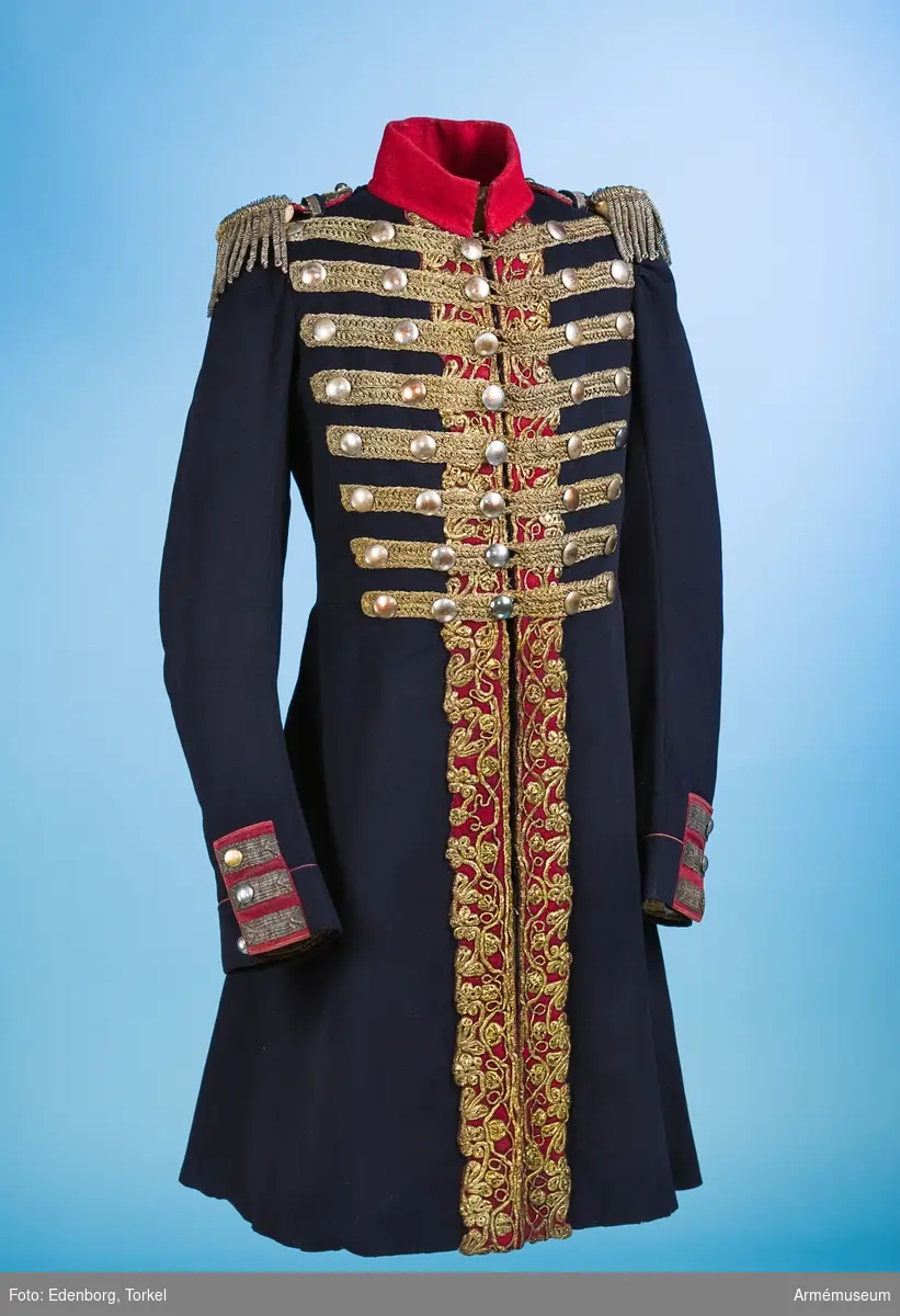Blå knälång rock med röd krage och snörmakerier av förgylld metalltråd.
Renoverad och försedd med nya ärmuppslag omkring 1831.

Rocken är sydd av fint mörkblått kläde och har snörmakerier av guldlan fastsydda på en remsa av rött kläde på båda sidor om knäppningen mitt fram, samt i slitsen bak. Dessutom revärer över bröstet utförda av förgyllda tvinnade metallsnoddar, och 5 rader med 8 rent dekorativa knappar i varje rad. Knapparna är släta och svagt välvda, och av en försilvrad kopparlegering. Den egentliga knäppningen är 2 x 7 hyskor och hakar kant i kant mitt fram, samt 2 x 2 dito i kragen.
Rocken är konstruerad med fram- och sidstycken i ett, samt helt bakstycke utan söm mitt bak. Den har midjesöm med separat skört. Skörtet har fram- och sidstycken i ett, och två smalare bakstycken vid slitsen bak. Röd ståndkrage av fint kläde, fodrad med det samma.
Ärmarna är formade tvåsömsärmar med fasta ärmuppslag med smal röd passpoal och röd matta med tre "knapphål" av metallgalon som pryds av tre mindre knappar.
De svängda sidsömmarna bak är dekorerade med samma förgyllda tvinnade metallsnoddar som revärerna. Vid ryggslutet sitter två knappar.
Bålen är fodrad med gråvitt fint ylletyg, medan ärmarna är fodrade med oblekt eller vitt linne.

Till rocken hör ett par epåletter av silvergalon med fransar av krusade och vridna silvertrådar.