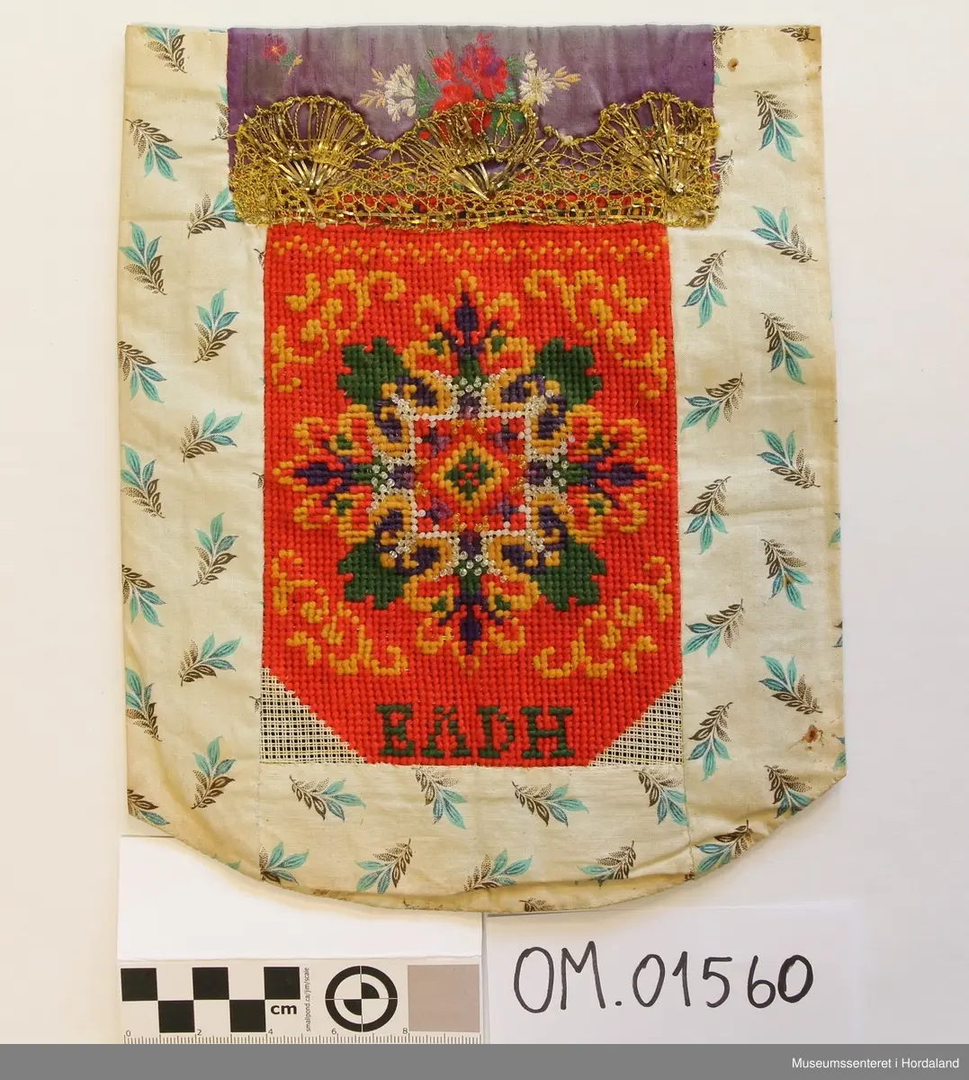 Bringeduk brodert med kross-sting på stramei i raudt, gult, grønt, lilla og kvite, blanke og gullfarga glasperler. Initialer er brodert nederst på broderiet. Blomstrete silkeband øvst. Kronekvarde i gull er montert eit stykke ned på broderiet og oppå silkebandet. Broderiet er montert på eit mønstrete bakstykke som og er brukt på kantane og under på framsida.