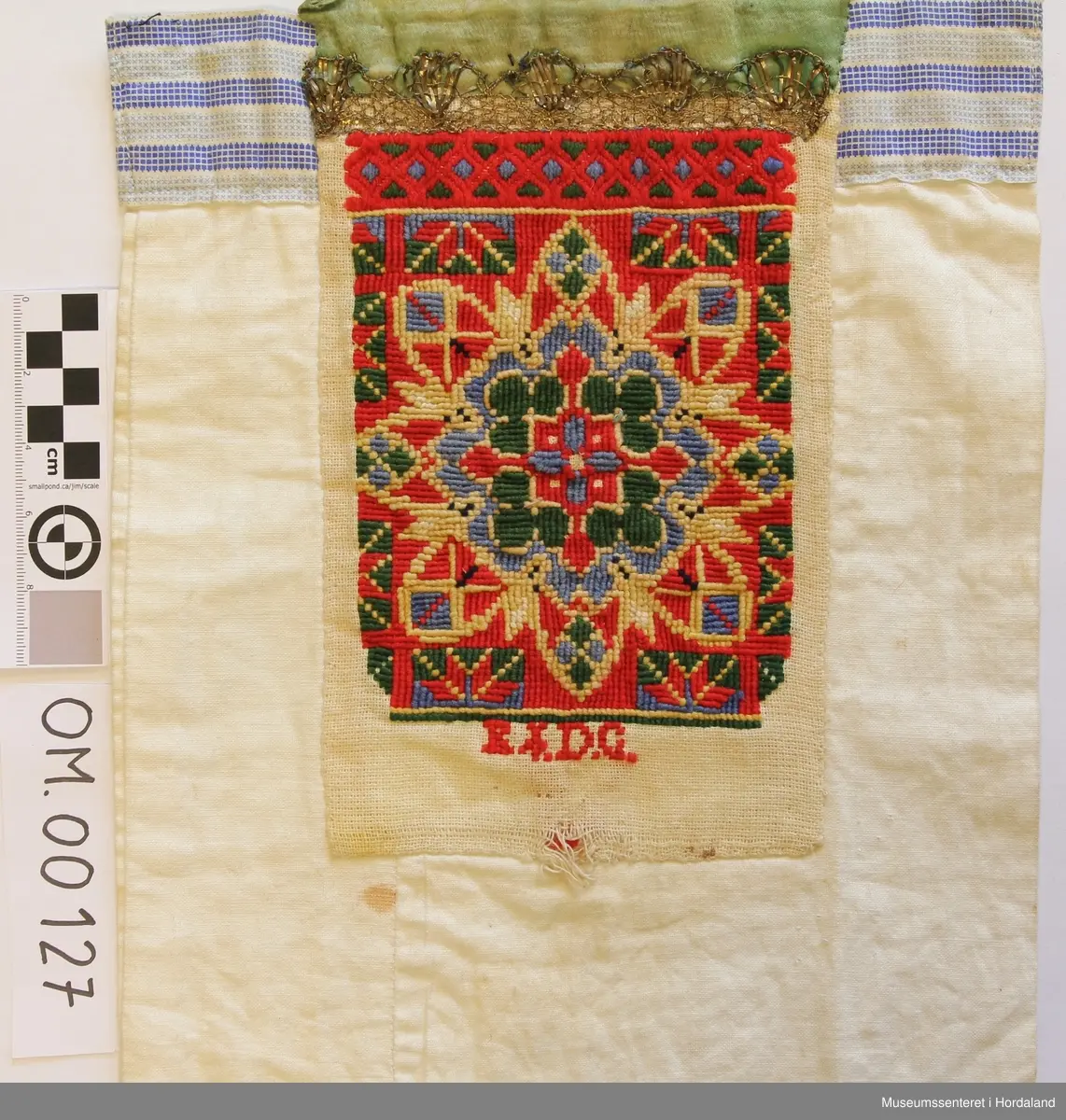 Bringeklut med broderi i fleire farger sydd på eit naturfarga stoff. Breit, grønt silkeband øvst. Gullfarga kronekvarde sydd oppå. Bakstykket er eit dobbelt kvitt stoff med ein kant oppe av eit mønstra stoff i striper, maskinsydd.
Under kronekvarden er det dragesaum. Ellers
kross-sting i broderiet.