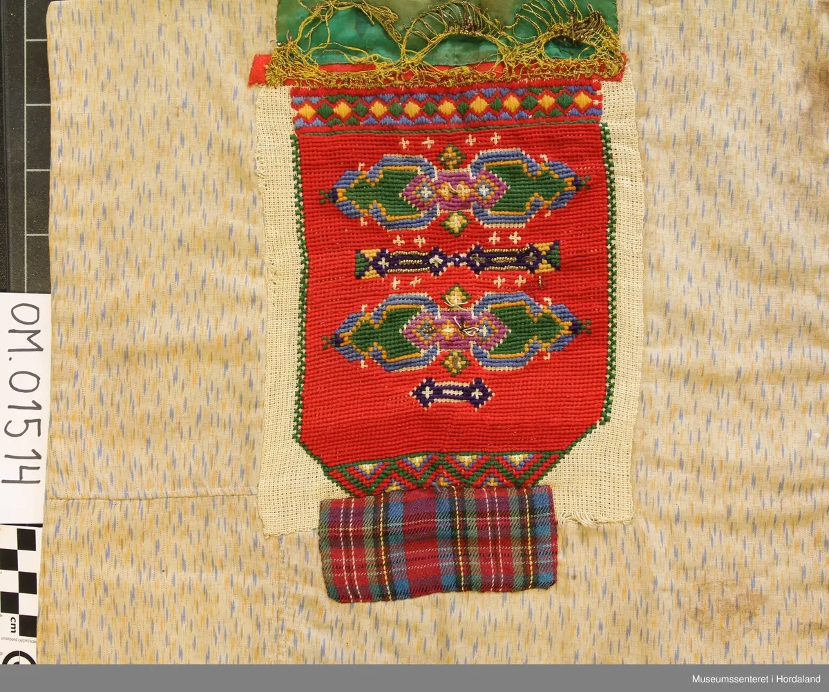 Bringeklut brodert i raudt med blå, grøn, gul, grøn, kvit, rosa og lilla. Gullfarga perler til dekor nokre stader, mange er knuste.  Grøn silkekvarde øvst med gullband i metall. Dette er sydd oppå eit raudt, smalt band. Rutete stoffbit sydd på under nederste broderte rad (Truleg på grunn av djup utringing på drakt). Montert på eit naturfarga, mønstra klede.