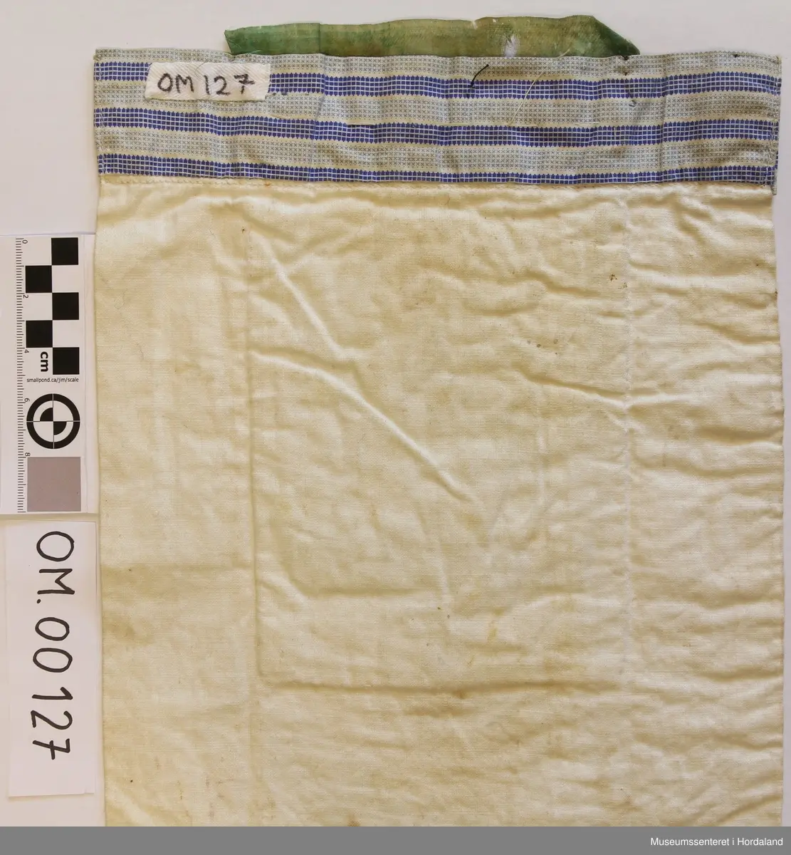 Bringeklut med broderi i fleire farger sydd på eit naturfarga stoff. Breit, grønt silkeband øvst. Gullfarga kronekvarde sydd oppå. Bakstykket er eit dobbelt kvitt stoff med ein kant oppe av eit mønstra stoff i striper, maskinsydd.
Under kronekvarden er det dragesaum. Ellers
kross-sting i broderiet.