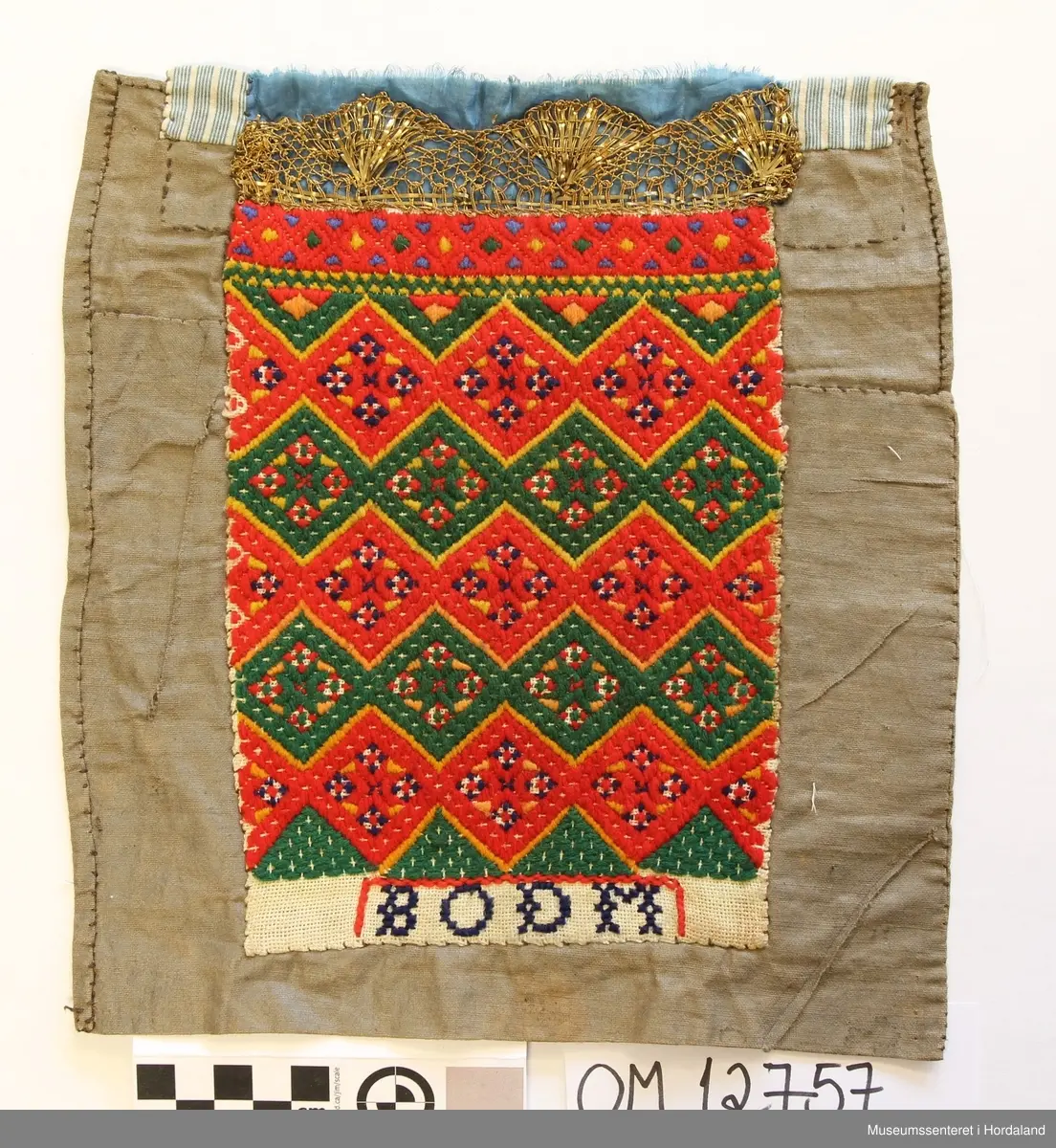 Brodert med ullgarn i plattsaum. Alt er handlaga. Hovudmotiv er små åttebladroser i border. Oppe er det påsydd eit blått silkeband med eit breitt gullband (i forgylt sølvtråd) utanpå silkebandet. Alt er montert på eit tynt, vokset bomullslerret. Oppe på baksida er det eit ekstra bomullstoff (blåstripet). Bokstavane BODM er broderte inn nede på bringeduken i kross-sting.