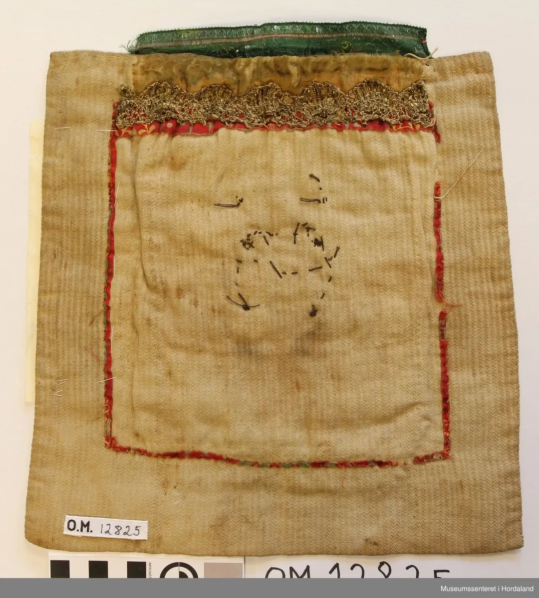 Bringeduk i ullbroderi med eit hovudmotiv i ei slags avrunda form som minner om eit granateple. I kvart hjørne er det små åttebladroser. I midten er det sydd på nokre store gullperler. Oppe er det brodert ei smal borde i plattsaum. Over den igjen er det eit breitt silkeband. Nede er det sydd på monogram : K H D E 
Sydd på eit kvitt teljestoff. Alt dette er montert på eit dobbelt, litt kraftig bomullstoff. 
På baksida av dette bomullstoffet viser det tydelege rester etter ein eldre bringeduk som har truleg bare vore eit mønstrete bomullstoff. Ovanfor dette stoffet er det ei kniplingsblonde i gull, montert utanpå eit silkeband som er så slitt at ein ikkje kan sei noko bestemt om kva farge det har vore.