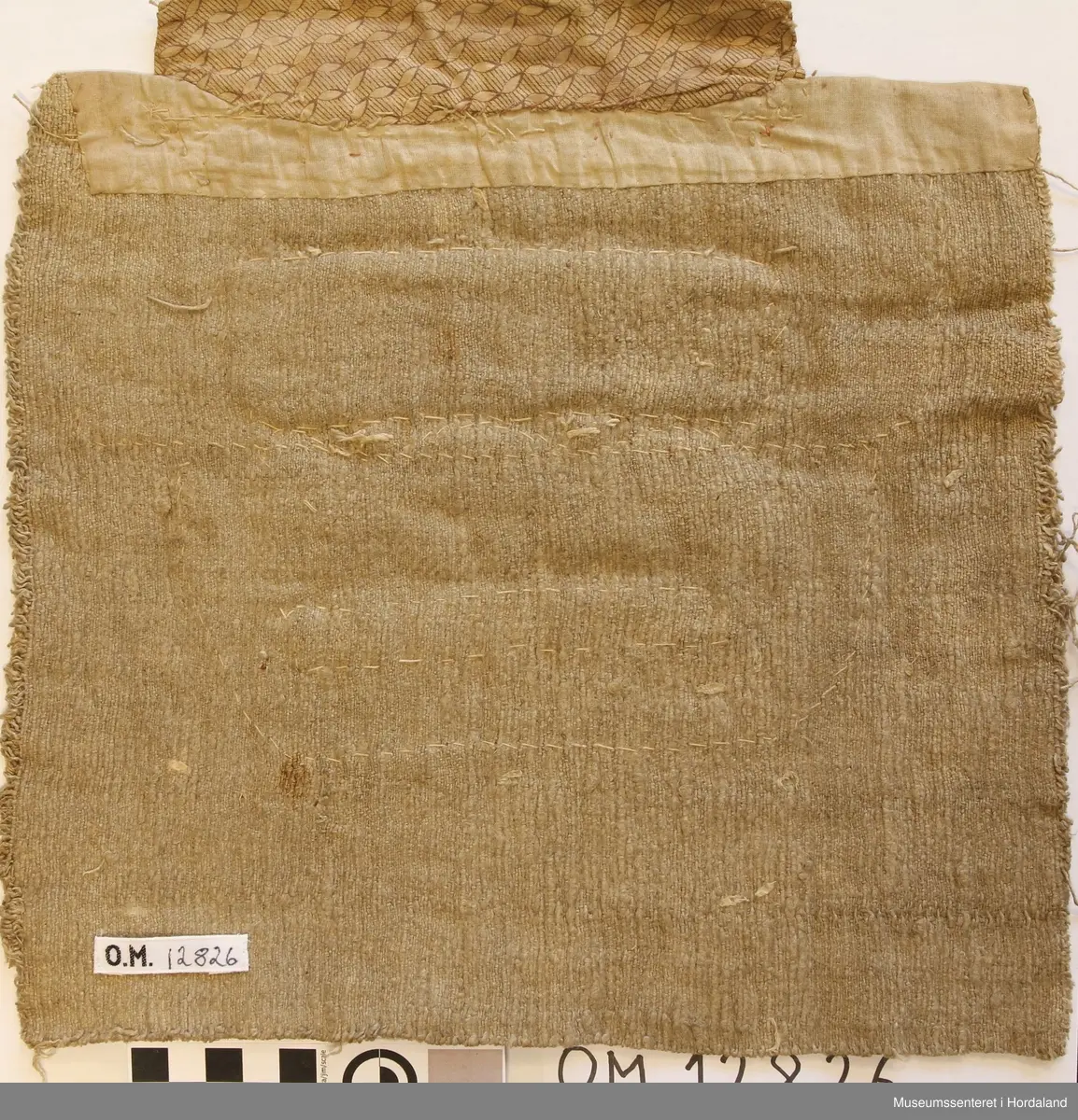 Bringeduk med eit perlenett forma som ei borde oppe og rett nedafor den er det to litt store, blanke glassteinar, så kjem ei brei metalborde i knipling og eit smalare vevd band i metall. Alt dette er montert på raudt bomullsflanell. Bakstykket er eit stykke grovt heimevove linstoff. Opp er bakstykket kanta med eit smalt stykke bomullslerret. Utanpå/ovanfor der att er det påsydd eit brunmønstra bomullstoff.