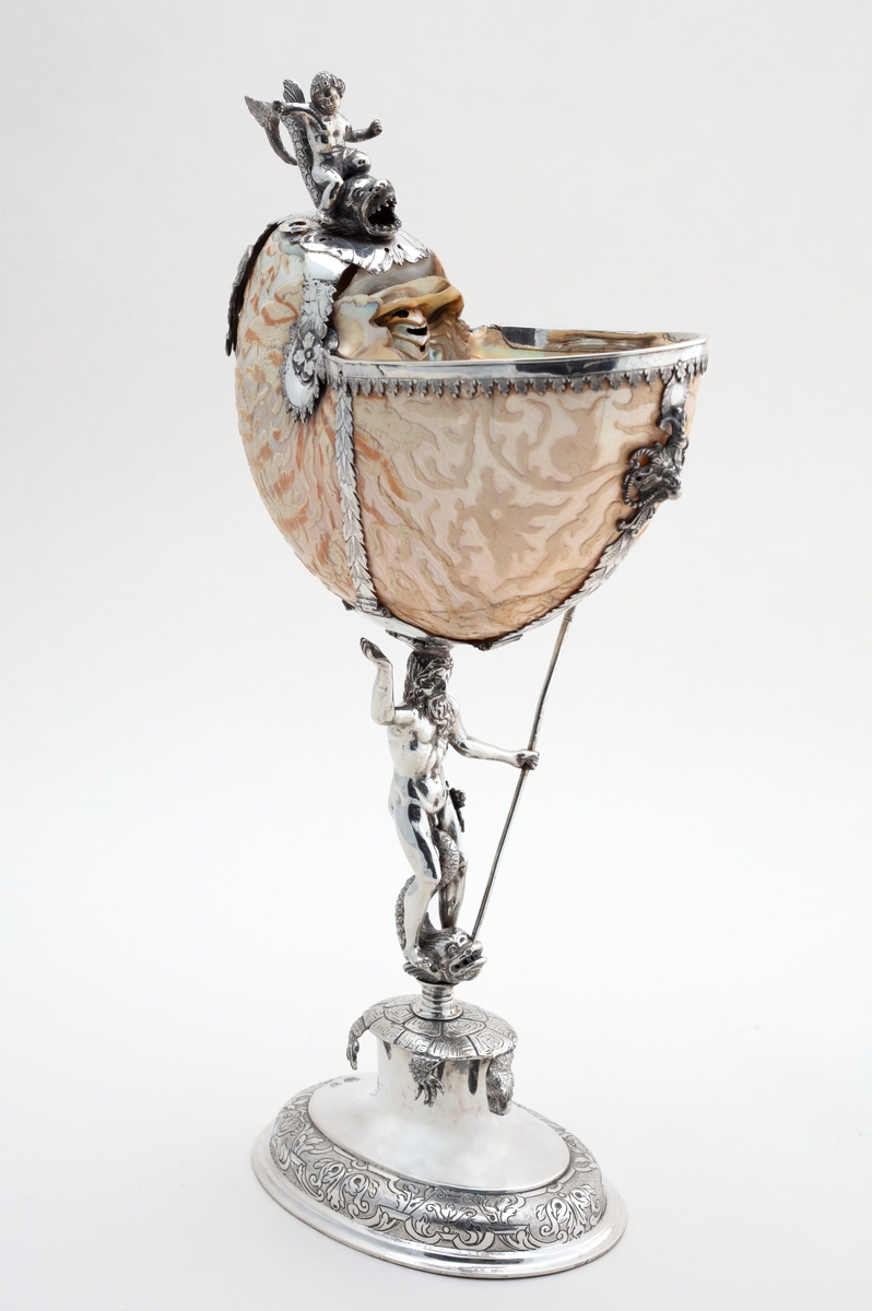 Skall av blekksprutarten Nautilus. Skåret dekorasjon, fot og innfatning sølv, støpt og gravert dekorasjon. Stpl: MF/H//1720 Morten Finchenhagen, Christiania. Skallet er brunflammet i overflaten. Nautilusblekkspruten lever i sydlige hav omkring Filippinene.