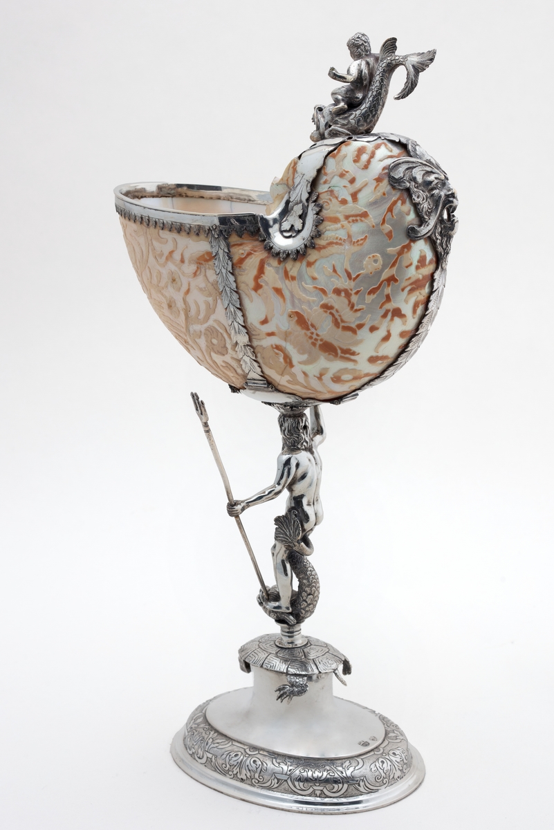 Skall av blekksprutarten Nautilus. Skåret dekorasjon, fot og innfatning sølv, støpt og gravert dekorasjon. Stpl: MF/H//1720 Morten Finchenhagen, Christiania. Skallet er brunflammet i overflaten. Nautilusblekkspruten lever i sydlige hav omkring Filippinene.