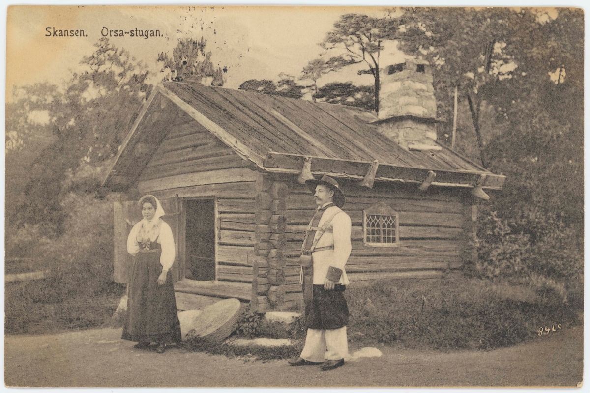 Vykort med motiv från Skansen. "Orsa-stugan". Man och kvinna i folkdräkt utanför stugan.