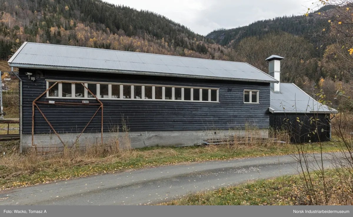 28. Smie og traktorstall, Mæl.
Smie og traktorstall på Mæl består av en eldre smie i vest og en i 1964 påbygd stall for jernbanetraktor/
robel i øst. Bygget er samlet 19x6 meter, smia er 6x6 meter og traktorstallen 13x6 meter. Det har betongfundament
og saltak tekt med bølgeblikk. Traktorstallen har liggende supanel og smia stående låvepanel,
alt beiset. Langsidene til traktorstallen har lange, smale og sammenhengende vindusrekker med 16 vinduer.
På nordfasaden er det dessuten et tofagsvindu. Østfasaden har tofløyet port med skråpanel og beslag for
innkjøring. Vestfasaden har en enfløyet låvepanelt dør. Sørfasaden har en tofløyet port med skråpanel inn
til smia, en fyllingsdør med fire speil og en dør med låvepanel. Interiøret i traktørstallen består av skinner
og grav for reparasjoner, og har liggende impregnert furupanel i vegger og tak.
Bygningen fremstår som autentisk og med integritet. Den har tilstandsgrad 1, jf. NS 3423.