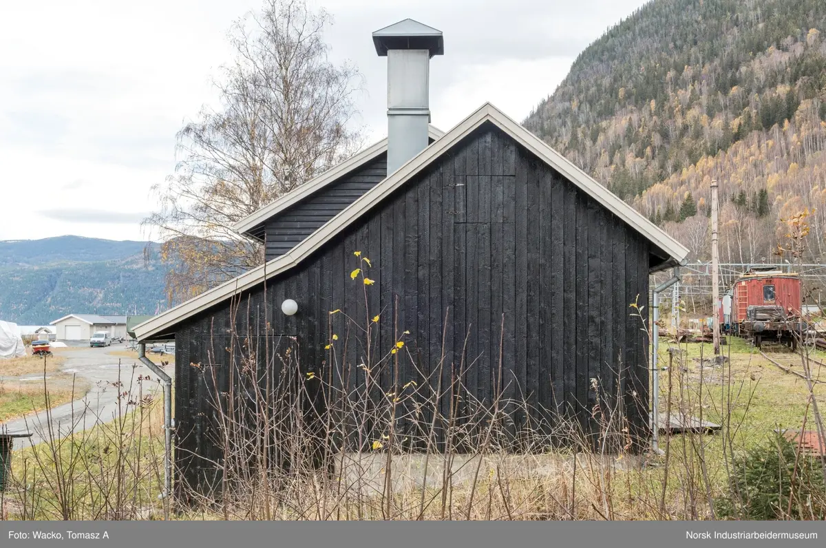 28. Smie og traktorstall, Mæl.
Smie og traktorstall på Mæl består av en eldre smie i vest og en i 1964 påbygd stall for jernbanetraktor/
robel i øst. Bygget er samlet 19x6 meter, smia er 6x6 meter og traktorstallen 13x6 meter. Det har betongfundament
og saltak tekt med bølgeblikk. Traktorstallen har liggende supanel og smia stående låvepanel,
alt beiset. Langsidene til traktorstallen har lange, smale og sammenhengende vindusrekker med 16 vinduer.
På nordfasaden er det dessuten et tofagsvindu. Østfasaden har tofløyet port med skråpanel og beslag for
innkjøring. Vestfasaden har en enfløyet låvepanelt dør. Sørfasaden har en tofløyet port med skråpanel inn
til smia, en fyllingsdør med fire speil og en dør med låvepanel. Interiøret i traktørstallen består av skinner
og grav for reparasjoner, og har liggende impregnert furupanel i vegger og tak.
Bygningen fremstår som autentisk og med integritet. Den har tilstandsgrad 1, jf. NS 3423.
