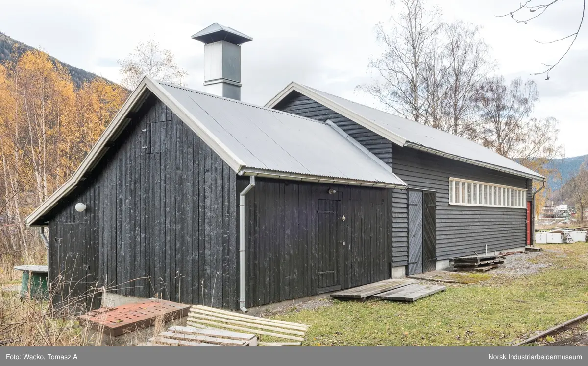28. Smie og traktorstall, Mæl.
Smie og traktorstall på Mæl består av en eldre smie i vest og en i 1964 påbygd stall for jernbanetraktor/
robel i øst. Bygget er samlet 19x6 meter, smia er 6x6 meter og traktorstallen 13x6 meter. Det har betongfundament
og saltak tekt med bølgeblikk. Traktorstallen har liggende supanel og smia stående låvepanel,
alt beiset. Langsidene til traktorstallen har lange, smale og sammenhengende vindusrekker med 16 vinduer.
På nordfasaden er det dessuten et tofagsvindu. Østfasaden har tofløyet port med skråpanel og beslag for
innkjøring. Vestfasaden har en enfløyet låvepanelt dør. Sørfasaden har en tofløyet port med skråpanel inn
til smia, en fyllingsdør med fire speil og en dør med låvepanel. Interiøret i traktørstallen består av skinner
og grav for reparasjoner, og har liggende impregnert furupanel i vegger og tak.
Bygningen fremstår som autentisk og med integritet. Den har tilstandsgrad 1, jf. NS 3423.