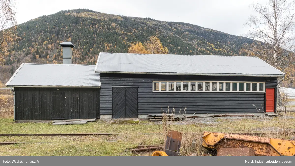 28. Smie og traktorstall, Mæl.
Smie og traktorstall på Mæl består av en eldre smie i vest og en i 1964 påbygd stall for jernbanetraktor/
robel i øst. Bygget er samlet 19x6 meter, smia er 6x6 meter og traktorstallen 13x6 meter. Det har betongfundament
og saltak tekt med bølgeblikk. Traktorstallen har liggende supanel og smia stående låvepanel,
alt beiset. Langsidene til traktorstallen har lange, smale og sammenhengende vindusrekker med 16 vinduer.
På nordfasaden er det dessuten et tofagsvindu. Østfasaden har tofløyet port med skråpanel og beslag for
innkjøring. Vestfasaden har en enfløyet låvepanelt dør. Sørfasaden har en tofløyet port med skråpanel inn
til smia, en fyllingsdør med fire speil og en dør med låvepanel. Interiøret i traktørstallen består av skinner
og grav for reparasjoner, og har liggende impregnert furupanel i vegger og tak.
Bygningen fremstår som autentisk og med integritet. Den har tilstandsgrad 1, jf. NS 3423.
