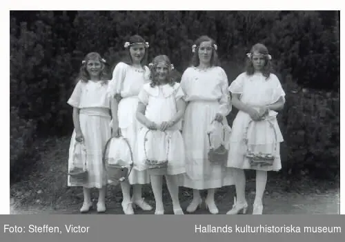 Troligen döttrar till anställda vid Fagereds sanatorium som är uppklädda och utsmyckade för att fira midsommar. (Doktor Steffen hade 9 döttrar så några kan vara hans flickor.) Bild 1: 5 flickor i vita klänningar och tidstypiska pannband med blommor på var sida, håller i blomstersmyckade korgar, alla olika. Bild 2: 10 flickor står i togadräkter i en talldunge med lågt sittande skärp och huvudkransar av blader, kanske lingon.