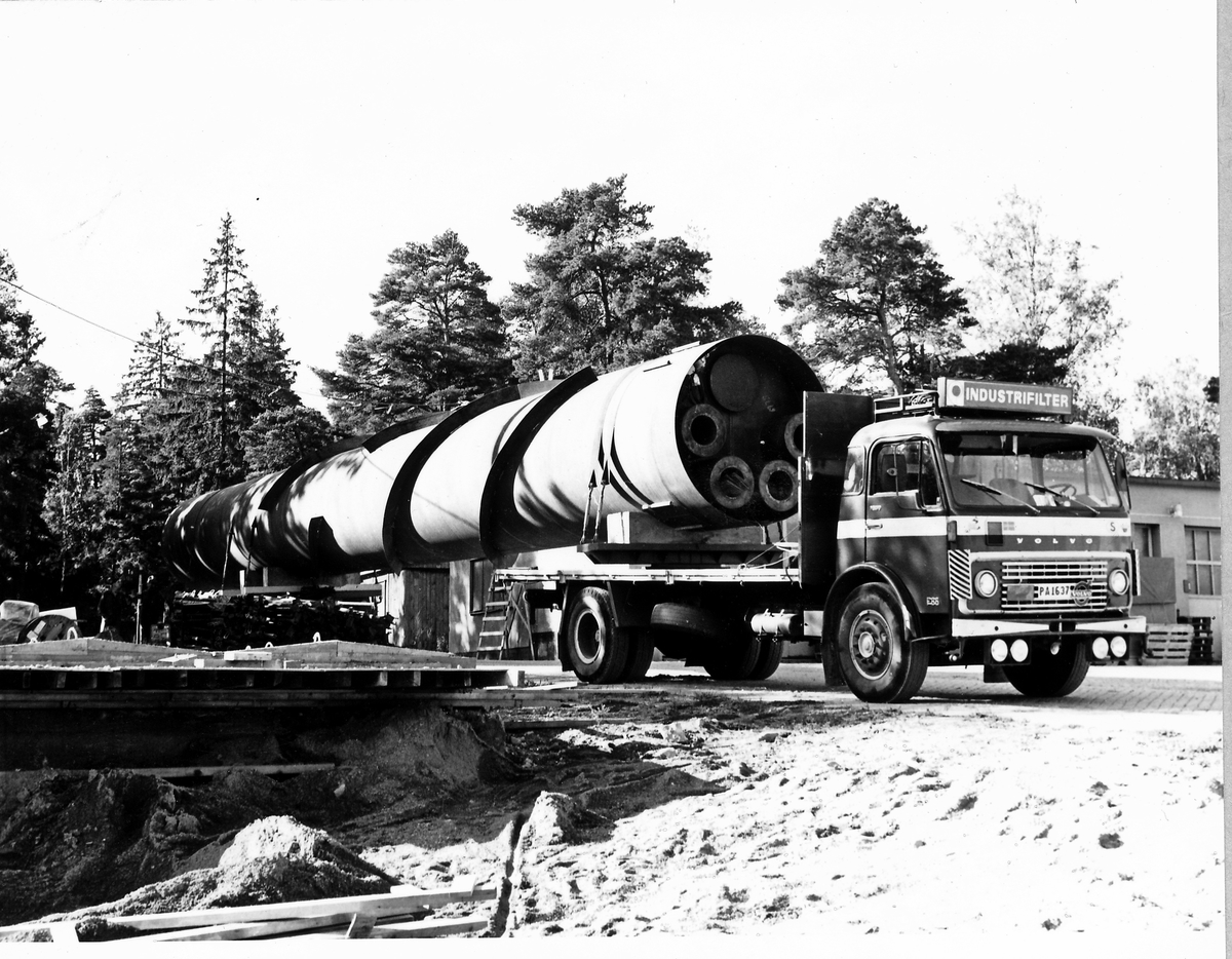 Montering av skorsten på regementets nya värmecentral den 10 oktober 1972.
Obs. Tio bilder.
Bild 1: Fundamentet på vilken den stora skorstenen skall skruvas fast.
Bild 2: Övre halvan av skorstenen under förflyttning till sin monteringsplats.
Bild 3: Nedre halvan av skorstenen håller på att lastas av från lastbilen. I bakgrunden syns den nya byggnaden till maskincentralen.
Bild 4: Nedre halvan läggs ned på stöd.
Bild 5: Den övre halvan av skorstenen, på yttersidan syns påsvetsade plåtspiraler för vindstabiliseringen.
Bild 6: Det krävdes kraftiga kranar för att klara alla lyft. I bakgrunden syns by 10 (Sörmlandssalen, markan och ubefmäss)
Bild 7: Flera kranar i samverkan för att få den nedre skorstenen i rätt läge.
Bild 8: Nu är skorstenen hopsatt och lyftet påbörjat.
Bild 9: Lyftet fortsätter, i förgrunden dom två oljecisternerna.
Bild 10: Nu står skorstenen på plats.