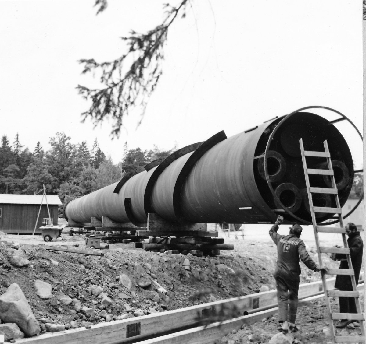 Montering av skorsten på regementets nya värmecentral den 10 oktober 1972.
Obs. Tio bilder.
Bild 1: Fundamentet på vilken den stora skorstenen skall skruvas fast.
Bild 2: Övre halvan av skorstenen under förflyttning till sin monteringsplats.
Bild 3: Nedre halvan av skorstenen håller på att lastas av från lastbilen. I bakgrunden syns den nya byggnaden till maskincentralen.
Bild 4: Nedre halvan läggs ned på stöd.
Bild 5: Den övre halvan av skorstenen, på yttersidan syns påsvetsade plåtspiraler för vindstabiliseringen.
Bild 6: Det krävdes kraftiga kranar för att klara alla lyft. I bakgrunden syns by 10 (Sörmlandssalen, markan och ubefmäss)
Bild 7: Flera kranar i samverkan för att få den nedre skorstenen i rätt läge.
Bild 8: Nu är skorstenen hopsatt och lyftet påbörjat.
Bild 9: Lyftet fortsätter, i förgrunden dom två oljecisternerna.
Bild 10: Nu står skorstenen på plats.