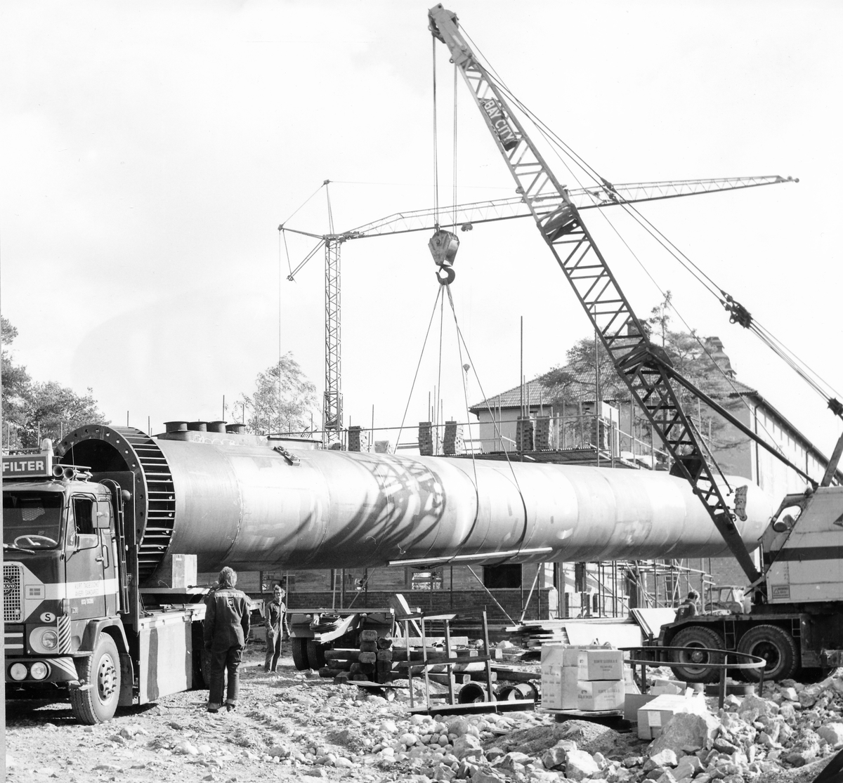 Montering av skorsten på regementets nya värmecentral den 10 oktober 1972.
Obs. Tio bilder.
Bild 1: Fundamentet på vilken den stora skorstenen skall skruvas fast.
Bild 2: Övre halvan av skorstenen under förflyttning till sin monteringsplats.
Bild 3: Nedre halvan av skorstenen håller på att lastas av från lastbilen. I bakgrunden syns den nya byggnaden till maskincentralen.
Bild 4: Nedre halvan läggs ned på stöd.
Bild 5: Den övre halvan av skorstenen, på yttersidan syns påsvetsade plåtspiraler för vindstabiliseringen.
Bild 6: Det krävdes kraftiga kranar för att klara alla lyft. I bakgrunden syns by 10 (Sörmlandssalen, markan och ubefmäss)
Bild 7: Flera kranar i samverkan för att få den nedre skorstenen i rätt läge.
Bild 8: Nu är skorstenen hopsatt och lyftet påbörjat.
Bild 9: Lyftet fortsätter, i förgrunden dom två oljecisternerna.
Bild 10: Nu står skorstenen på plats.