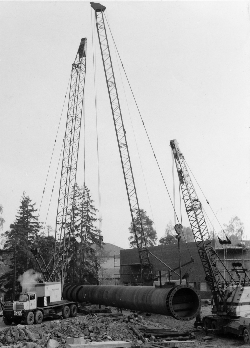 Montering av skorsten på regementets nya värmecentral den 10 oktober 1972.
Obs. Tio bilder.
Bild 1: Fundamentet på vilken den stora skorstenen skall skruvas fast.
Bild 2: Övre halvan av skorstenen under förflyttning till sin monteringsplats.
Bild 3: Nedre halvan av skorstenen håller på att lastas av från lastbilen. I bakgrunden syns den nya byggnaden till maskincentralen.
Bild 4: Nedre halvan läggs ned på stöd.
Bild 5: Den övre halvan av skorstenen, på yttersidan syns påsvetsade plåtspiraler för vindstabiliseringen.
Bild 6: Det krävdes kraftiga kranar för att klara alla lyft. I bakgrunden syns by 10 (Sörmlandssalen, markan och ubefmäss)
Bild 7: Flera kranar i samverkan för att få den nedre skorstenen i rätt läge.
Bild 8: Nu är skorstenen hopsatt och lyftet påbörjat.
Bild 9: Lyftet fortsätter, i förgrunden dom två oljecisternerna.
Bild 10: Nu står skorstenen på plats.