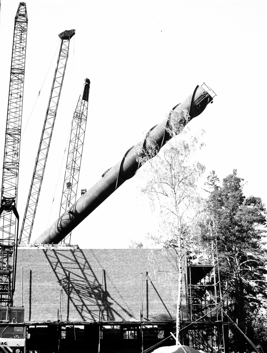 Montering av skorsten på regementets nya värmecentral den 10 oktober 1972.
Obs. Tio bilder.
Bild 1: Fundamentet på vilken den stora skorstenen skall skruvas fast.
Bild 2: Övre halvan av skorstenen under förflyttning till sin monteringsplats.
Bild 3: Nedre halvan av skorstenen håller på att lastas av från lastbilen. I bakgrunden syns den nya byggnaden till maskincentralen.
Bild 4: Nedre halvan läggs ned på stöd.
Bild 5: Den övre halvan av skorstenen, på yttersidan syns påsvetsade plåtspiraler för vindstabiliseringen.
Bild 6: Det krävdes kraftiga kranar för att klara alla lyft. I bakgrunden syns by 10 (Sörmlandssalen, markan och ubefmäss)
Bild 7: Flera kranar i samverkan för att få den nedre skorstenen i rätt läge.
Bild 8: Nu är skorstenen hopsatt och lyftet påbörjat.
Bild 9: Lyftet fortsätter, i förgrunden dom två oljecisternerna.
Bild 10: Nu står skorstenen på plats.