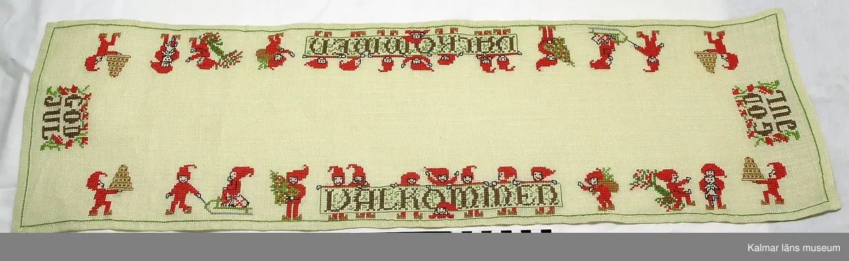 KLM 41657:6 Jullöpare, linne. Rektangulär, vitt tuskaftvävt linne med broderat mönster i korssöm i rött, brunt och grönt, små tomtar i olika scener samt texten VÄLKOMMEN och GOD JUL.