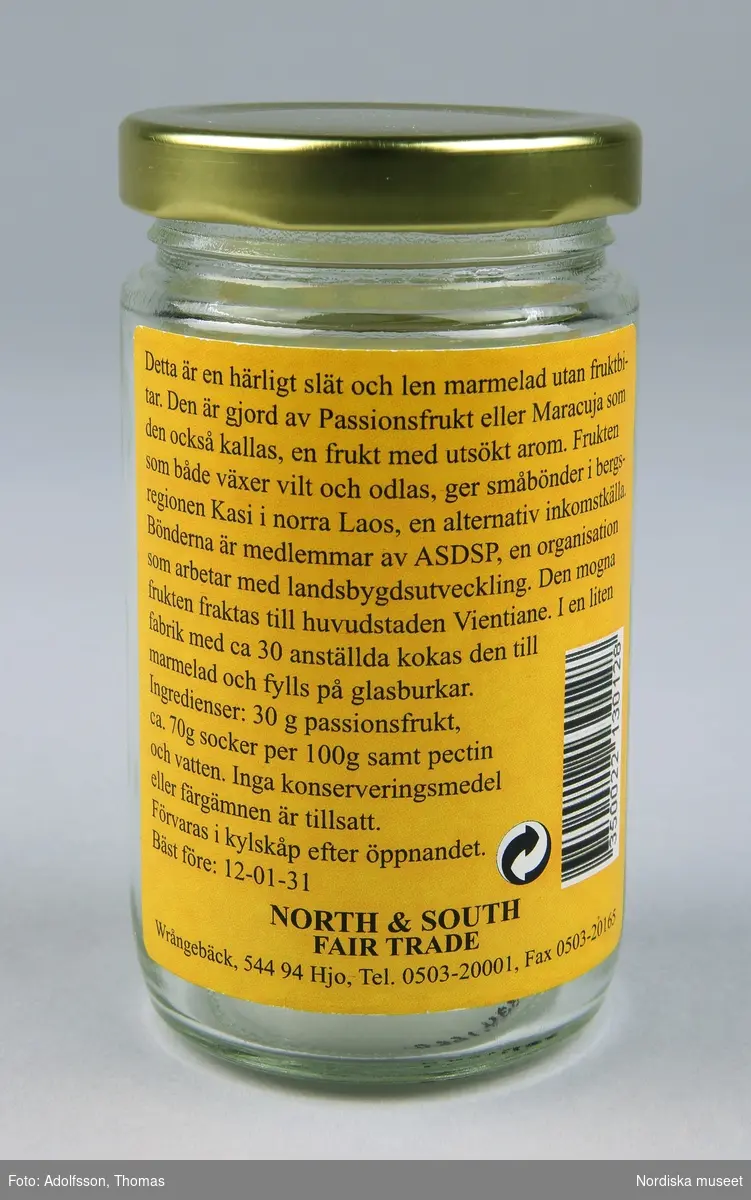 a) Marmeladburk med b) lock och påklistrade etiketter. På frontvinjetten: "North and South Fair Trade Passionsfrukt Marmelad från småbönder i Laos 275g".  Baksidesetiketten beskriver böndernas produktionsförhållanden samt fabriken i huvudstaden Vientiane där marmeladen kokas.