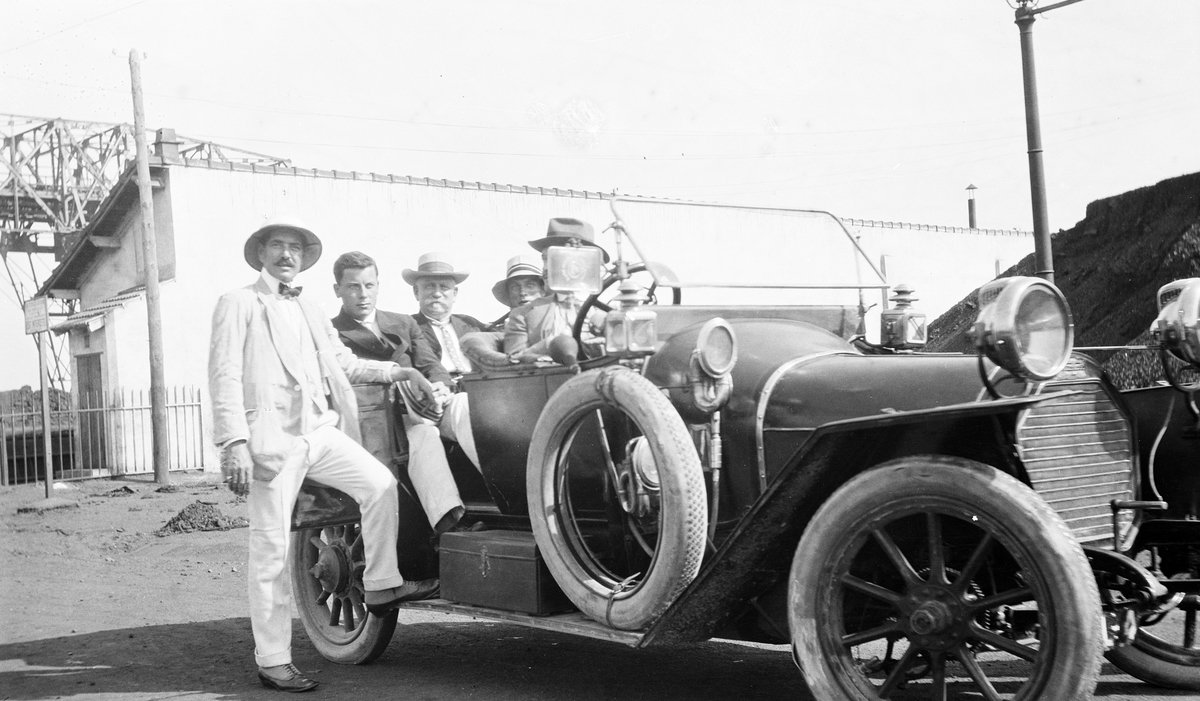 Åpen bil (den samme som ble fraktet med D/S STORFOND) på land. Bak i bilen sitter kaptein Gustav Waage mellom sønnene sine. Motivet er tatt i et havneområde.