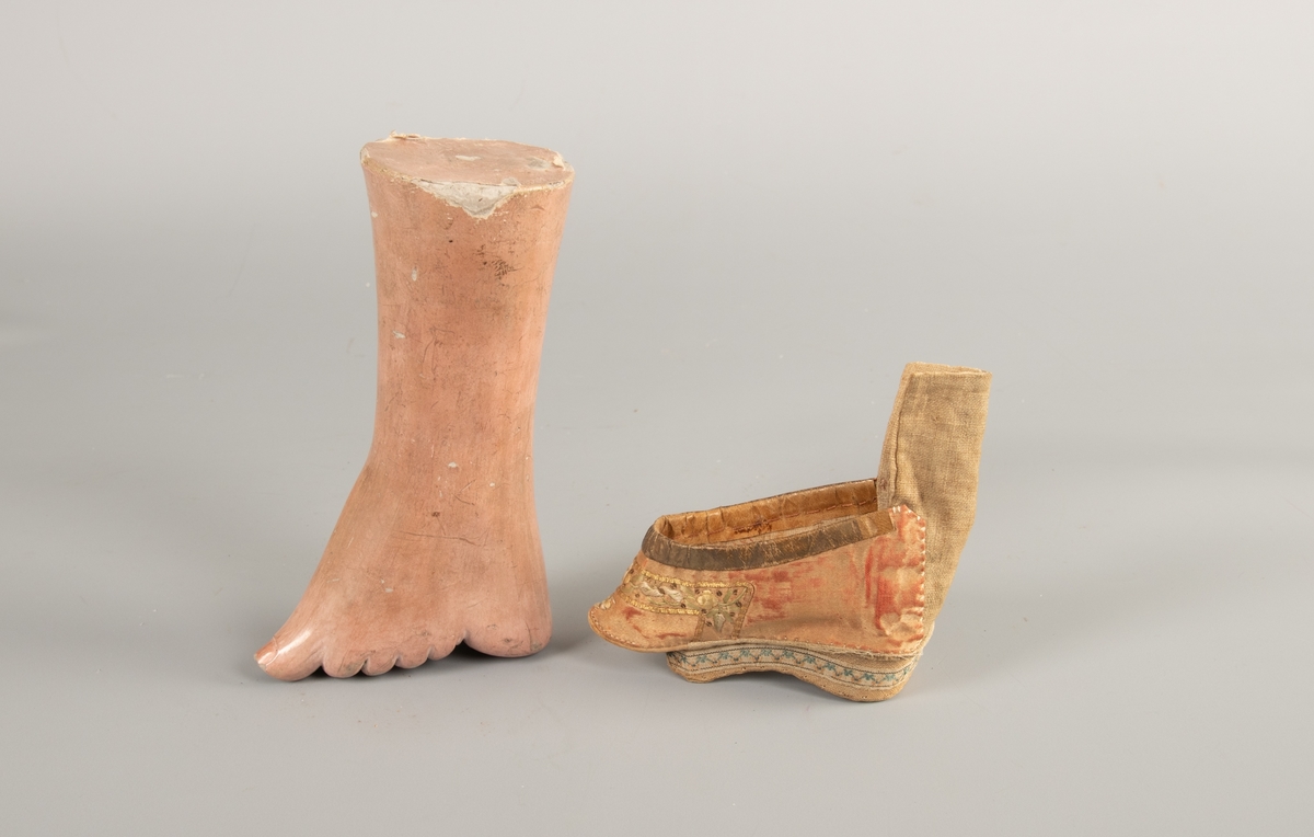 a) Fot i   gips  med malt   papp  utenpå. Sko:   silke og   lin. Hælen er sterkt forskjøvet, som en sokkel under skoen.  Tøffelen har en gyllen farge og et bredt   forgylt  bånd i overkant. Bunnfargen virker som om den opprinnelig har  vært sterkere og dypere rød.   Broderier  over tåen og langs kanten av hælen.
