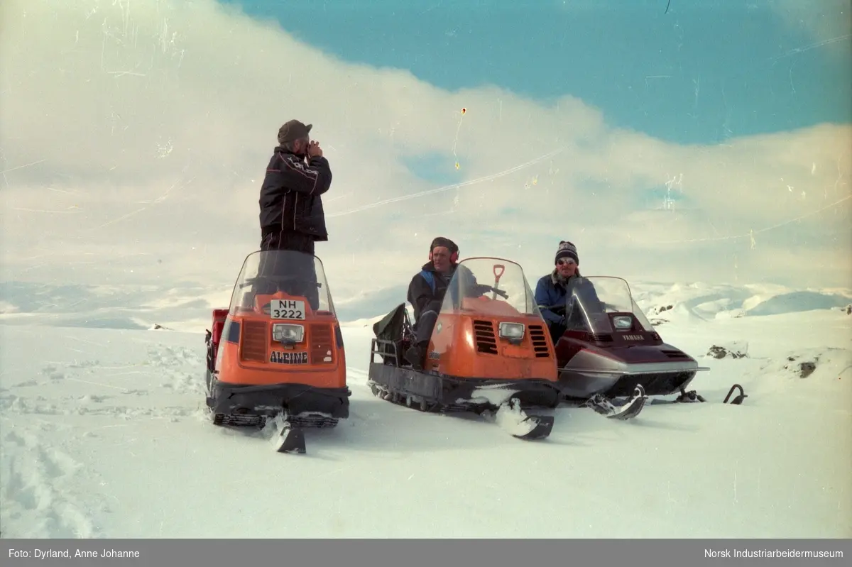 Tre menn på snøskuter i fjellet, antagelig Kvamsfjell på Møsstrond. Petter Hagen står på skuter og speider. Bjørn Dyrland med øreklokker og Olav O. Vågen med solbriller