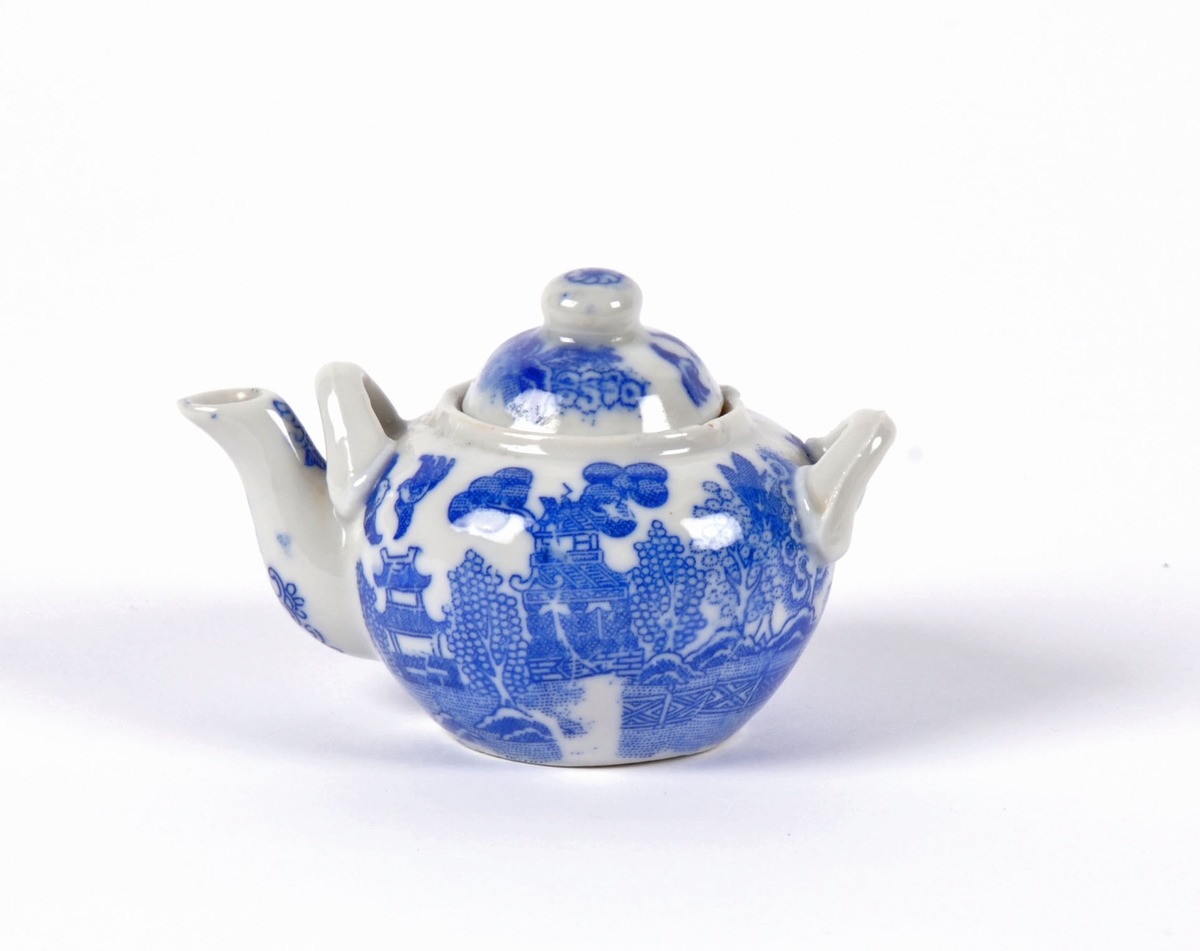Dukkeservise i 12 deler; 6 tekopper og kanne, 2 kaffekopper og kanne, fløtemugge og sukkerskål. Dekoren har mønster i blått og hvitt; det populære "willow pattern" eller piletremønsteret.