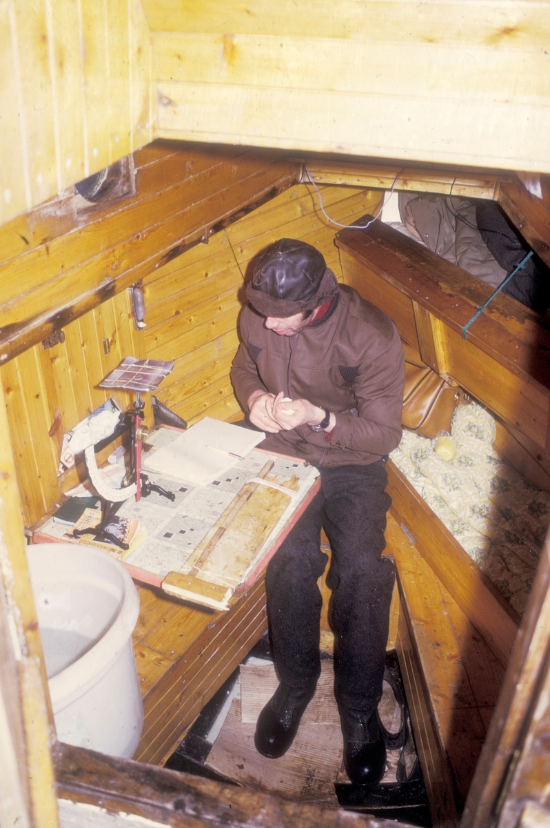 Flakstadvåg, 1973 : En mann sitter ved et bord i lugaren på en båt. På bordet foran seg har han ei vekt, et målebrett og en notatbok. Her taes det prøver av smolt.