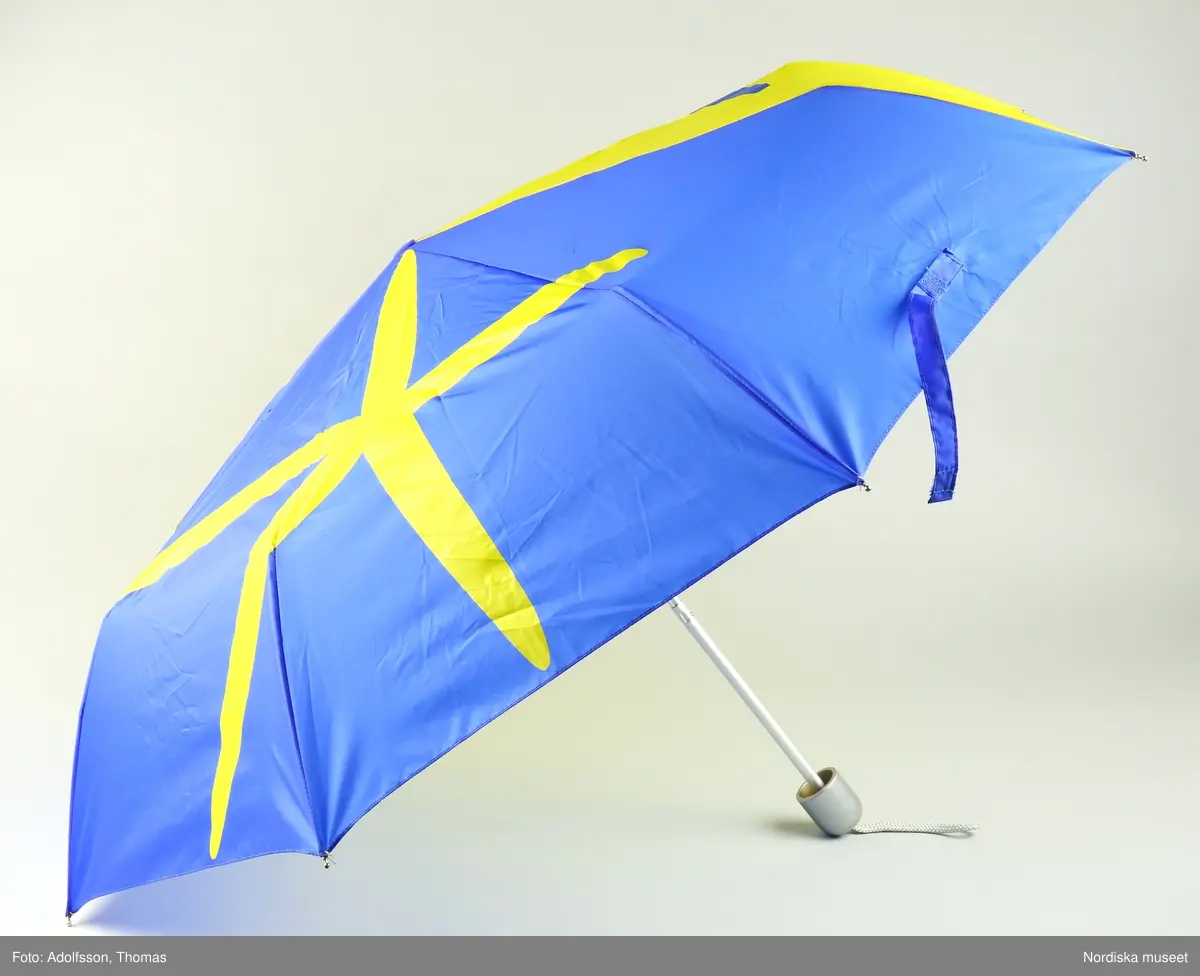 Hopfällbart paraply med stomme av vitmetall och plast, skaft av aluminium, silverfärgat handtag av trä med silverfärgad snodd. Paraplytyg av blå och gul nylon i EU med den officiella logotypen för Sveriges ordförandeskap 2001 och text ”eu2001.se”.  Blått band med kardborrelås för att hålla samman det hopfällda paraplyet. På undersidan en vidsydd lapp med text ”WACKES 033/206600”.
/Annika Tyrfelt 2013-06-27