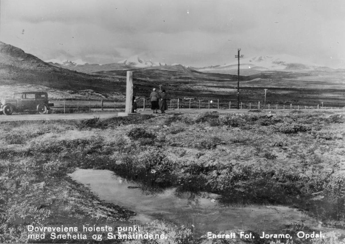 Dovrefjell, bil og folk ved merket av Dovrefjellsvegens høyeste punkt. Utsikt mot Snøhetta til høyre og Svånåtindan